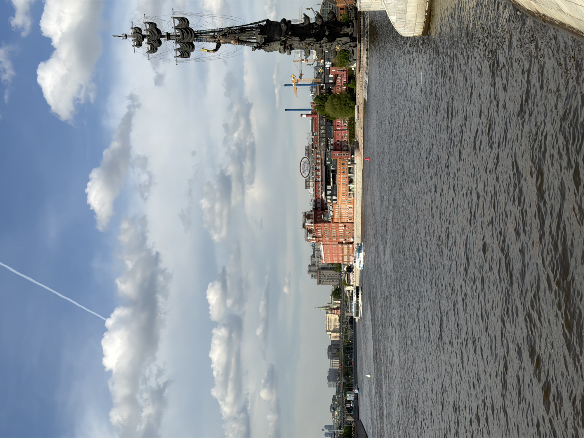 The image depicts a scene in Moscow, Russia. The shot seems to be taken from a high vantage point, possibly a bridge, looking along the Moskva River. On the left, there's a view of the city with historical buildings, including the "Red October" factory. The sky is filled with clouds. The Peter the Great Statue is visible on the left side as well. The river flows alongside the buildings, occupying the right portion of the picture.