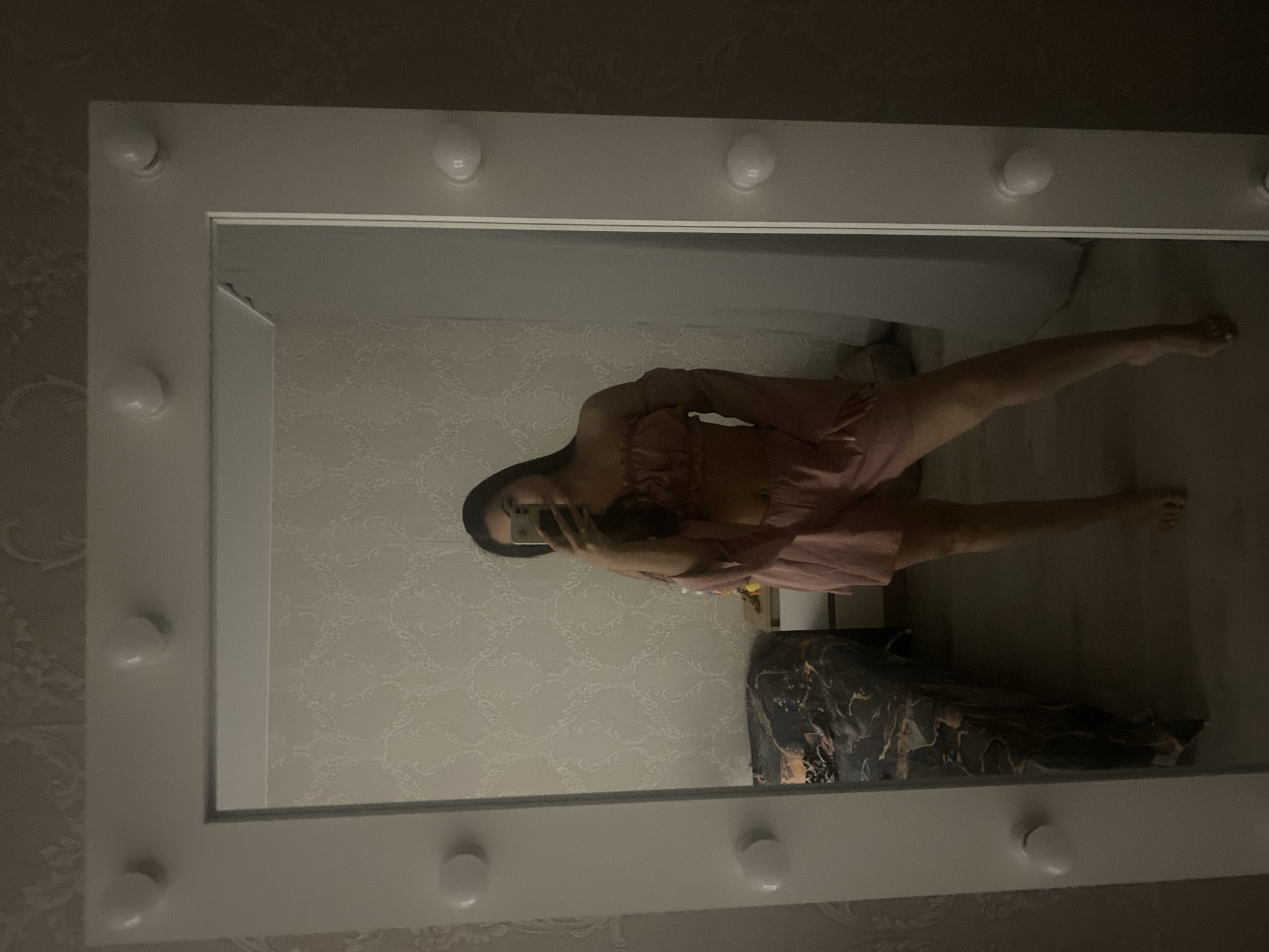 A young woman is taking a mirror selfie in a bedroom. She is wearing a pink off-the-shoulder crop top with bell sleeves and matching pink shorts. She is holding a smartphone in her right hand, angled to capture her reflection. The mirror is large and framed with lights. In the background, there is a bed with a patterned comforter, a nightstand, and textured wallpaper. The floor appears to be wood. The lighting is dim, with the bulbs in the mirror frame being the primary light source. The overall impression is of a casual, personal moment in a domestic setting. The image does not provide any information to determine its location in the city of Chaikovsky, Russia.