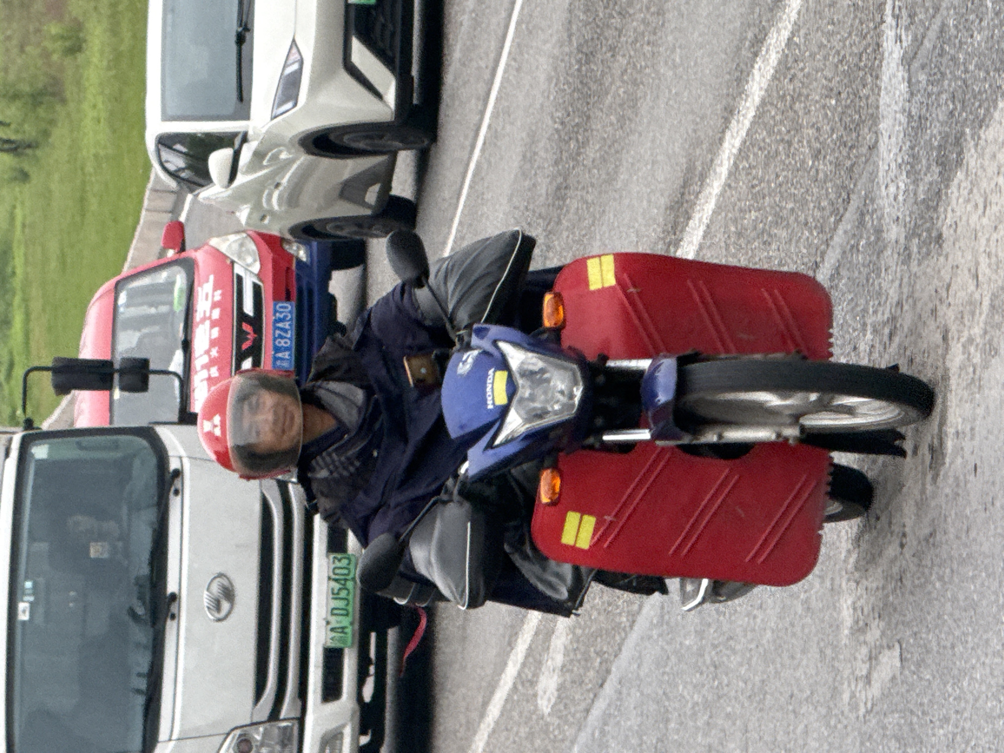 A man on a blue Honda motorcycle, equipped with red side panniers, is captured on a paved road in Jiangjin, China. The image is rotated 90 degrees counter-clockwise, showing the scene vertically.
The man is wearing a red helmet and a dark blue jacket, looking directly towards the camera with a neutral expression. The motorcycle has "HONDA" written on its front fairing, and the red panniers feature yellow reflective strips.
Other vehicles are visible around the motorcycle. To the man's left (forward in the direction of travel), there's a red truck or van with a partially visible license plate reading "A 8ZA30". Chinese characters are also visible on its side, possibly indicating the brand "金杯" (Jinbei). Further ahead of the red vehicle is a white truck with a green license plate displaying "A DJ5403". Behind the motorcycle, a white SUV or car can be seen.
The setting is an outdoor road with visible white lane markings. Green foliage or grass is present in the background, suggesting a roadside environment. The lighting indicates it is daytime, possibly overcast, and the road surface appears slightly damp or reflective. The vehicles seem to be in traffic or stopped momentarily.