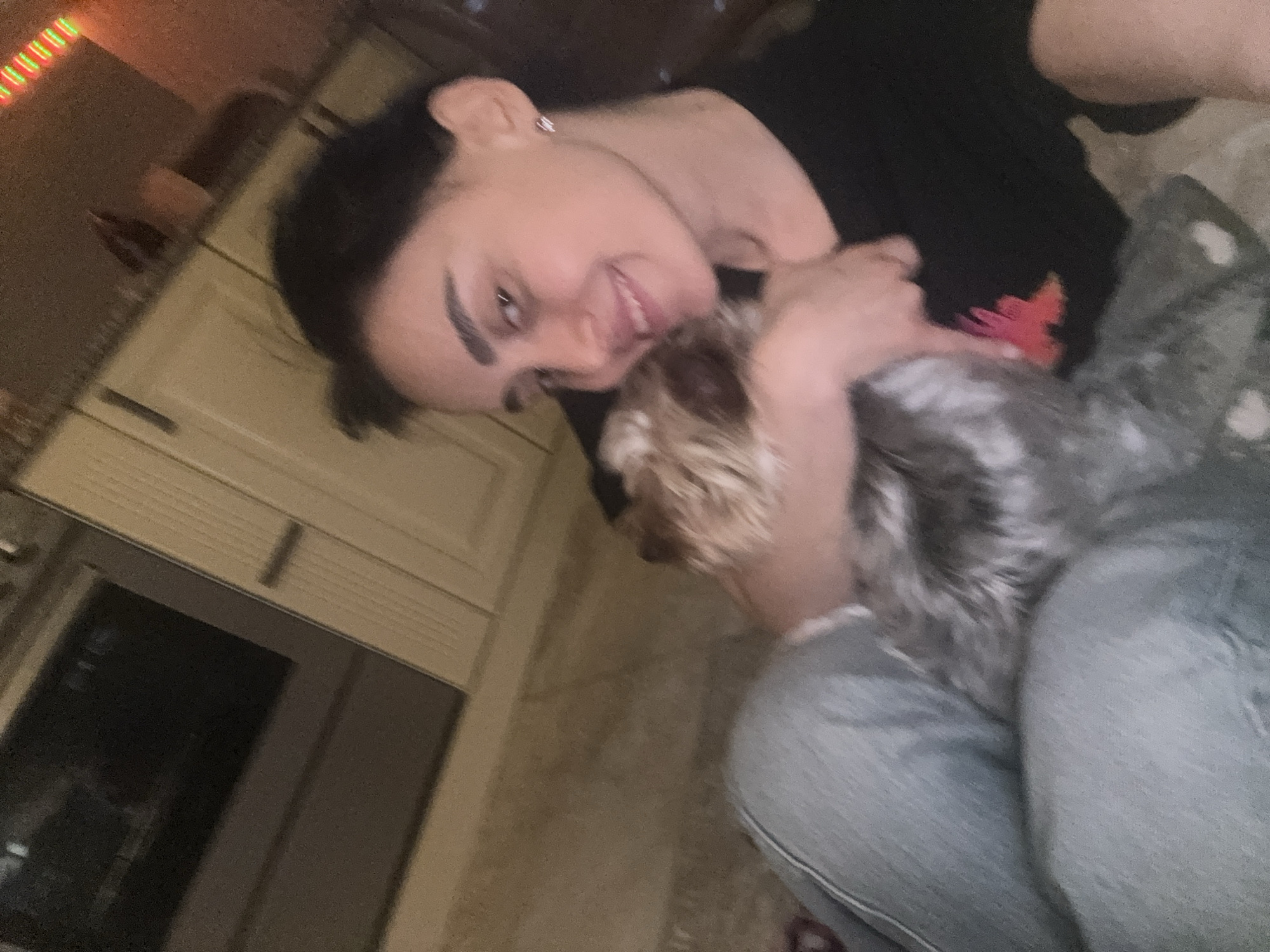 A woman is pictured indoors, cradling a small dog. She has dark hair pulled back, fair skin, and is wearing a dark top and light-colored jeans. The dog is light brown and gray. They appear to be in a kitchen, judging by the cabinets and appliances visible in the background. The image location may be Tbilisi, Georgia, based on the prompt, but there are no visual indicators in the image itself to confirm this location.