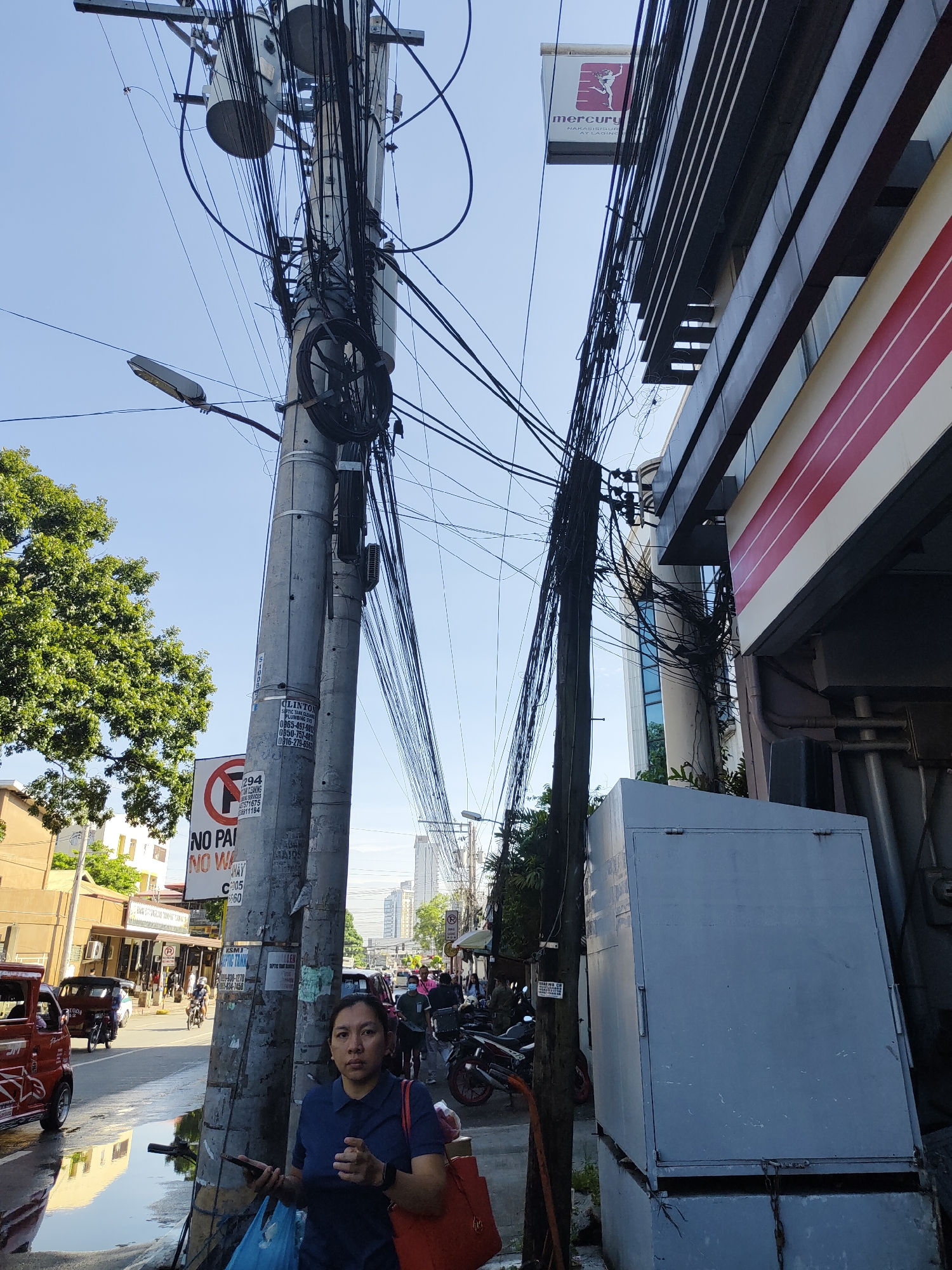 This full-shot street photograph captures a bustling urban scene in Davao City, Philippines, under clear daylight. In the foreground, a woman with dark hair, wearing a blue polo shirt and carrying an orange bag, stands next to a utility pole. She holds a smartphone in her left hand and looks directly at the camera with a neutral expression.
The scene is dominated by multiple utility poles and a dense, chaotic entanglement of overhead electrical and communication wires that crisscross the bright blue sky. A street light is attached to one pole. To the right, a large grey electrical box sits on a concrete base next to another wooden pole, also laden with wires. Beyond this, a modern building with horizontal red and white stripes and dark glass windows is visible, featuring a "Mercury Drug" sign with the text "NAKASISIGUR" and "Y LAGING".
In the background, a busy street is visible with various vehicles, including a red tricycle and motorcycles, along with several pedestrians. Buildings line the street. A prominent "NO PARKING NO WAITING" sign is attached to the main utility pole. Other visible text includes "CLINTON PLUMBING" and phone numbers such as "0865-497-9", "0950-757-481", "0916-279-6561", "294", "NG SERVICES", "7571675", "18911184", "C005", "KSMI", "938-1278", and "1965-834-7458". The bright sunlight indicates it is daytime.