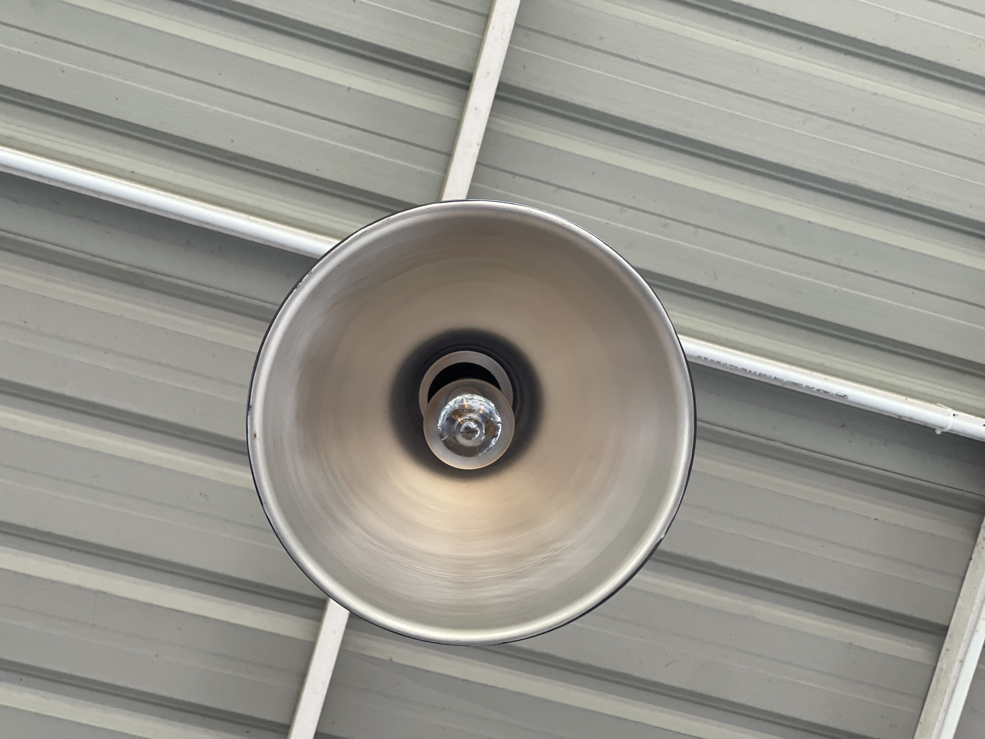 This is a close-up, downward-facing shot of a metallic pendant light fixture with a visible incandescent bulb. The light fixture is suspended from a ceiling made of corrugated metal, painted in a light gray color with alternating raised and recessed bands. Two white pipes or conduits run diagonally across the ceiling, one above and to the left of the light fixture, and another to the right and slightly below it. The image appears to be taken indoors, possibly in a commercial or industrial space, with no indication of the city of Pulokiton, Indonesia, or any people present. The lighting is diffuse, suggesting ambient light rather than direct sunlight.