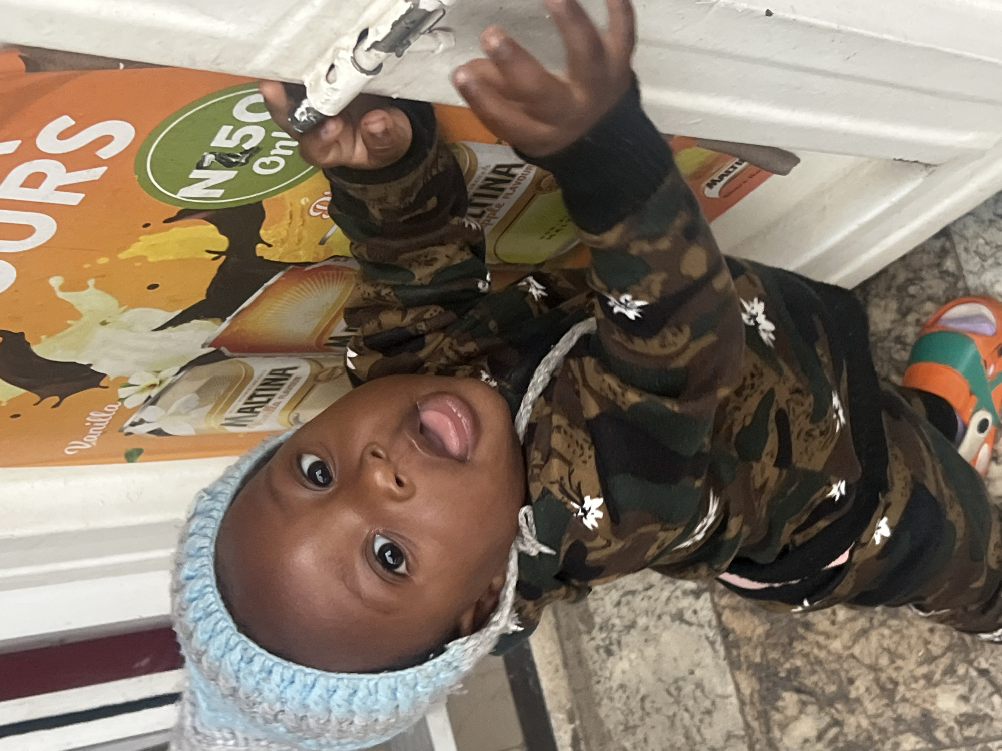 A toddler wearing a light blue knit cap and a camouflage-patterned jacket is gripping the edge of a shelf or display case. The toddler is facing the camera with their tongue sticking out. The shelf or display case has advertisements for "Maltina" Vanilla and Apple flavor drinks. A price tag of "N50 Only" is visible. The location is inferred to be Gura Top, Nigeria, based on the prompt. Part of an orange and green shoe is visible in the background, indicating the toddler is standing on a tiled or patterned floor.