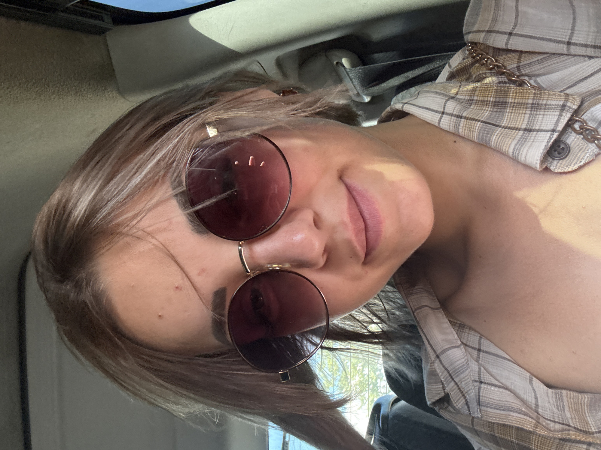 A young woman with light brown hair and sunglasses is sitting in a car. She is wearing a plaid shirt. The environment appears to be a sunny day, with sunlight illuminating parts of the scene. There are hints of the car's interior, such as the seatbelt and the ceiling. The image does not contain information to place it in Yuzhno-Sakhalinsk, Russia.