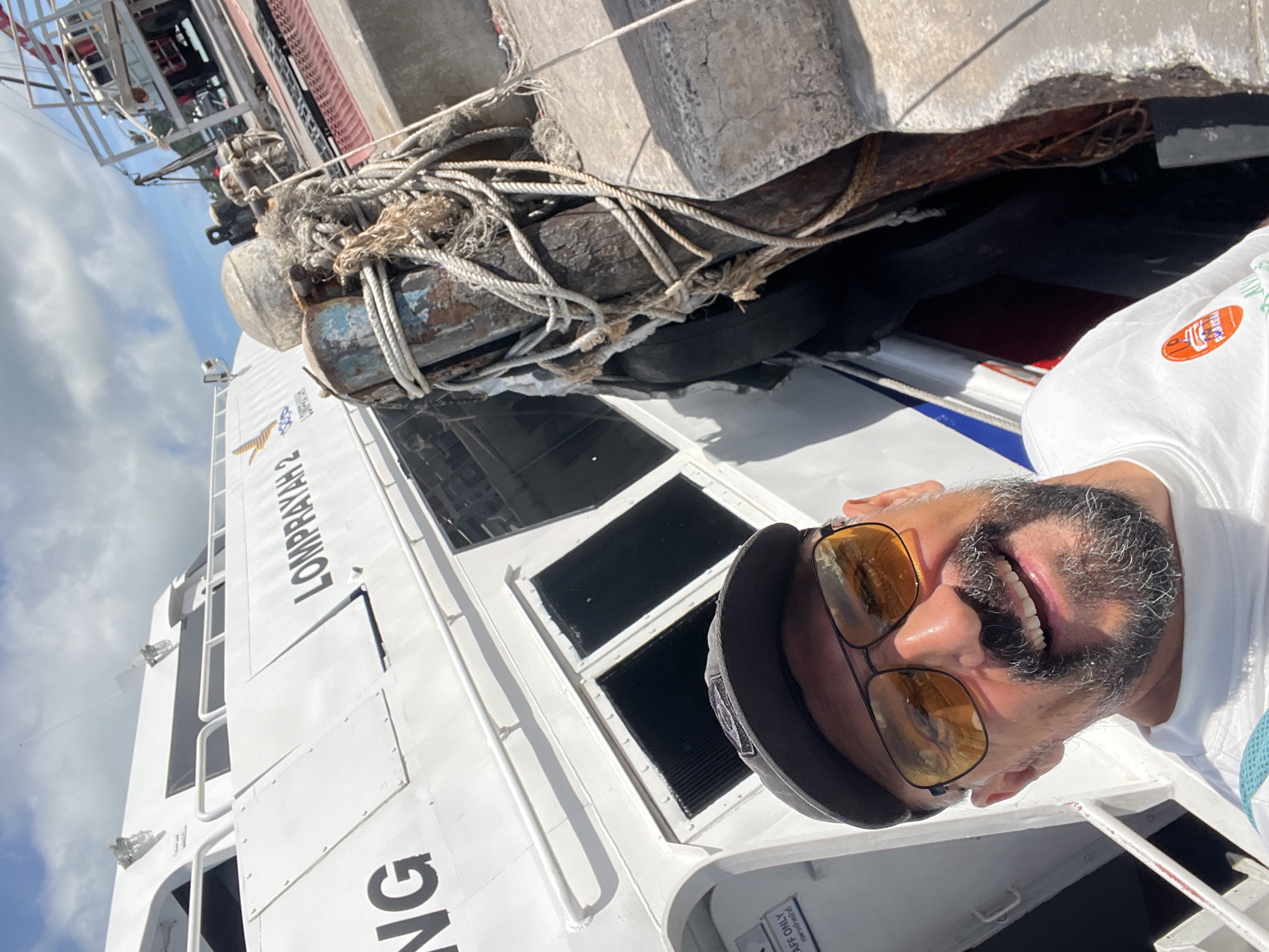 A man with sunglasses and a beard smiles at the camera, wearing a white t-shirt and a cap. He is standing next to a large white ferry named "LOMPRAYAH 2" docked at a pier in Koh Phangan, Thailand. Ropes and mooring bollards are visible on the pier. The sky is cloudy.