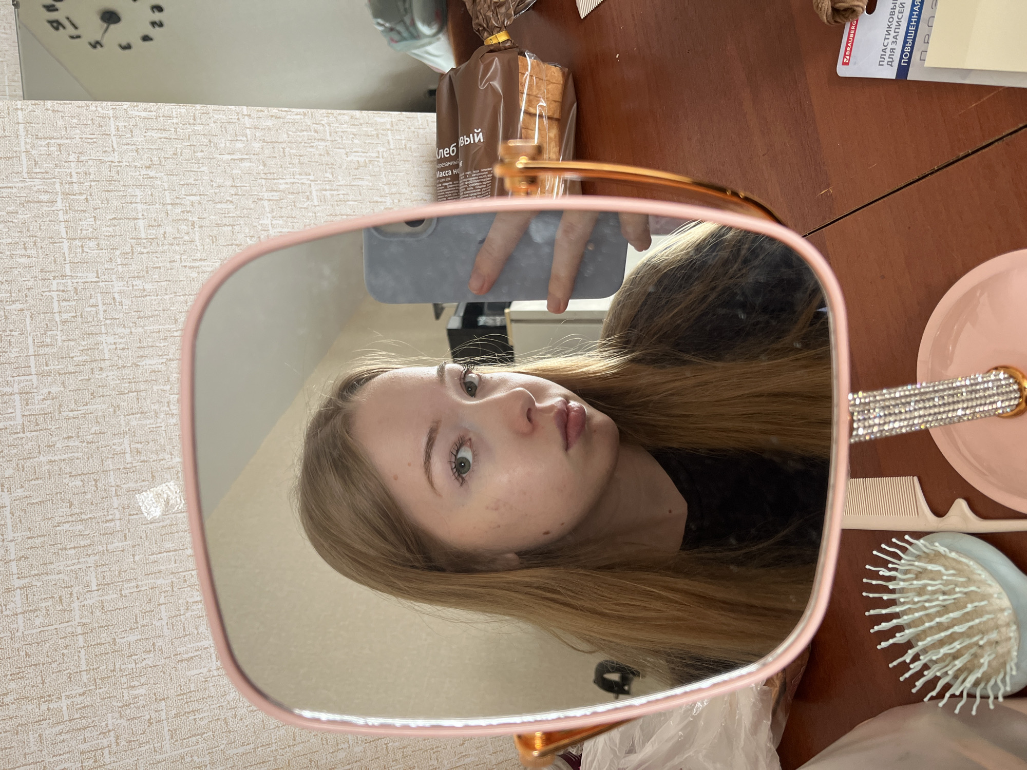 The image is a mirror selfie featuring a young woman with long, light brown hair and green eyes. She is captured in the reflection of a pink-framed, rose gold vanity mirror, holding a light grey smartphone that is taking the picture. Her face is slightly turned, and she looks directly into the lens with a neutral expression. Some freckles or small marks are visible on her face.
The setting is indoors, likely a personal room, indicated by a patterned light-colored wallpaper and a wooden table surface. On the table, outside the mirror's frame, are a light blue hairbrush with white bristles, a white comb, a pink circular dish, and a decorative object adorned with rhinestones.
Within the mirror's reflection, besides the woman and her phone, a clock is visible on the wall, showing hands positioned around 10 or 11 o'clock, suggesting a late morning time. A loaf of pre-sliced bread in a brown plastic package is also visible in the reflection.
Visible text on the bread package includes Russian words like "ХЛЕБНЫЙ" (bread/bready) and "нарезной" (sliced). A stationery label visible on the table features "V BRAUBERG" and "ПЛАСТИКОВЫЙ ДЛЯ ЗАПИСЕЙ" (plastic for notes) in Russian. These textual cues support the location context of Chaikovsky, Russia. The overall lighting is bright and natural, consistent with daytime.