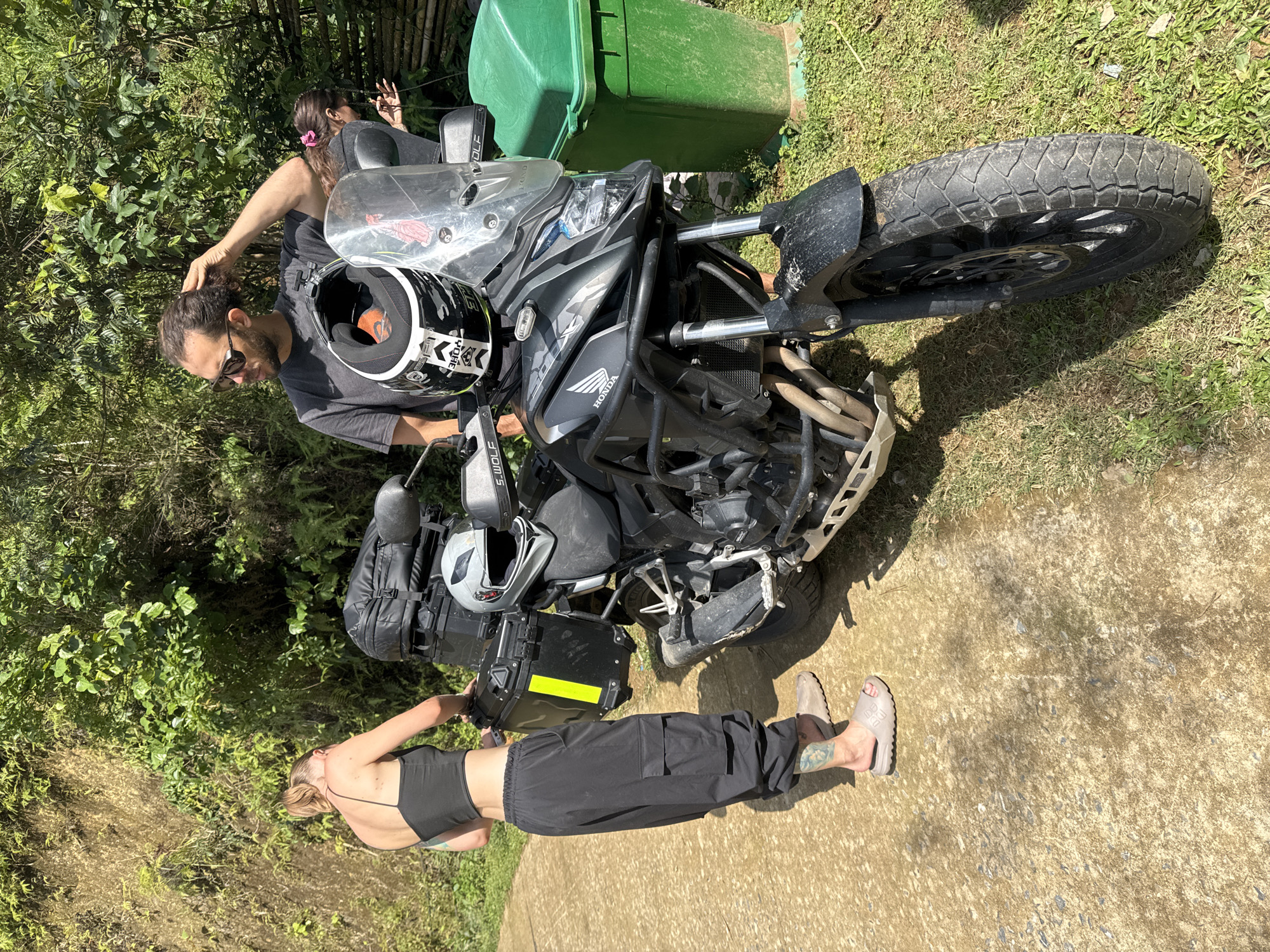 A medium shot captures a man and a woman alongside a Honda motorcycle in Ngã Ba Kim, Vietnam. The man, wearing sunglasses and a dark t-shirt, is touching his hair while standing by the motorcycle. The woman, clad in a black top and pants, stands with her hand on the motorcycle’s luggage. The motorcycle, laden with bags and helmets, is parked on a paved surface adjacent to a grassy area and lush greenery. A green garbage bin is visible behind the motorcycle.