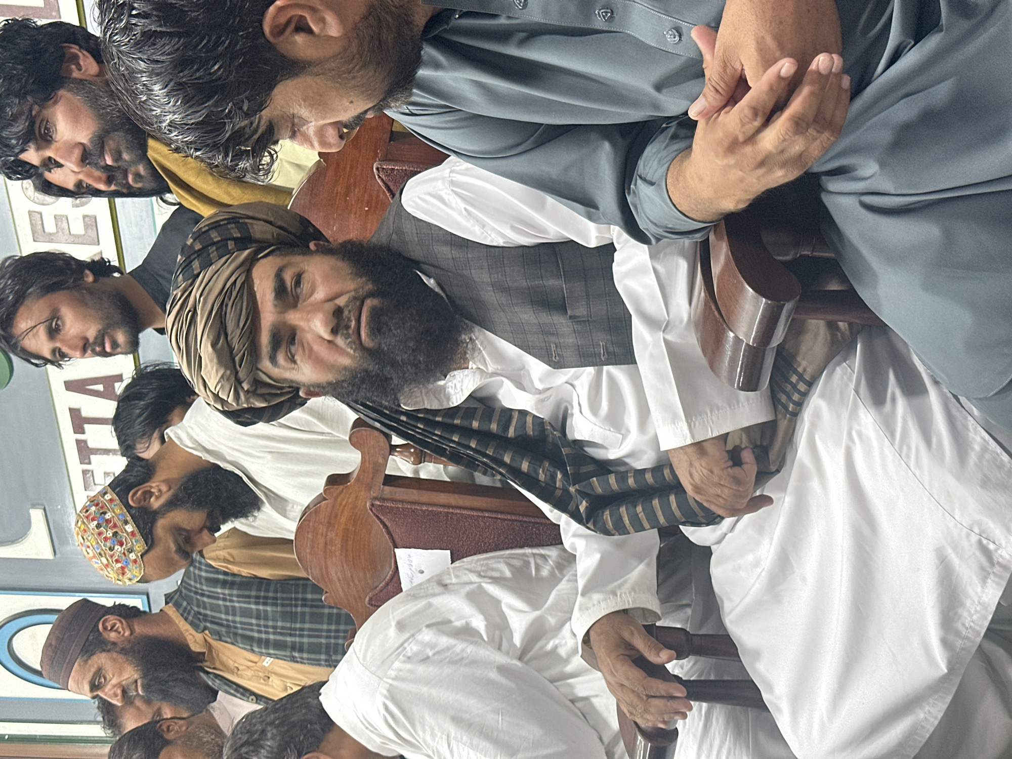 The image shows a group of men, most with beards and traditional clothing, seated in a room. A man in a light-colored turban and a vest sits prominently in the middle of the frame. The men are situated in chairs, and some are looking in different directions. The location is Quetta City Tehsil, Pakistan, as indicated by signage that is partially visible. The men appear to be in an indoor setting, possibly a gathering or meeting.