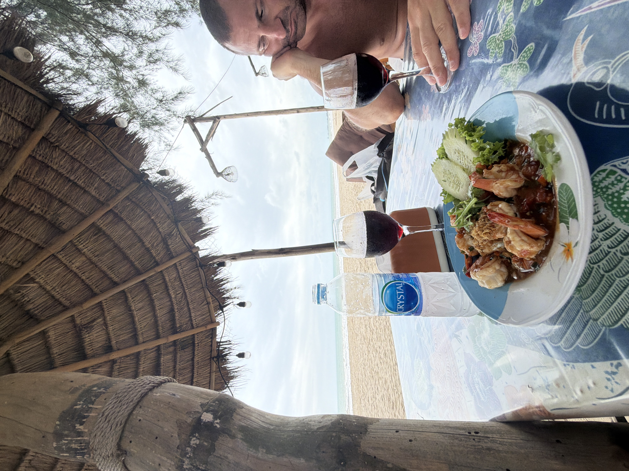 An outdoor beachfront restaurant in Bang Muang, Thailand, features a thatched roof providing shade. In the foreground, a table is set with a blue and white plate of shrimp and vegetables, two glasses of red wine, and a bottle of Crystal water. A man, shirtless, sits at the table, resting his head on his hand, looking towards the beach. The beach and ocean are visible in the background under a cloudy sky. A wooden pillar supporting the thatched roof is on the left side of the image.