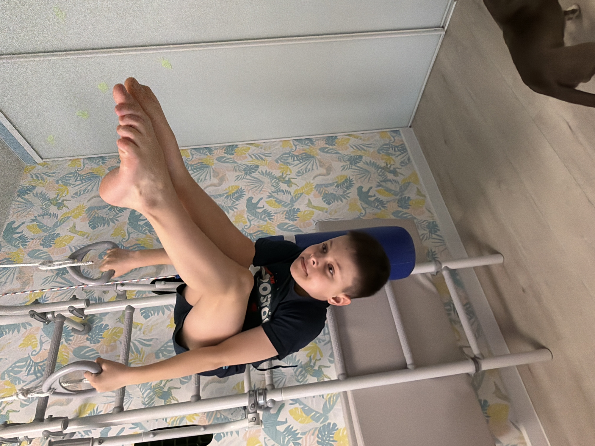A boy is hanging upside down on a gymnastic ladder with handles in a room with wallpaper featuring plants and animals. The room appears to be indoors, possibly in Votkinsk, Russia. The boy is holding onto the ladder with his hands and his legs are raised. A section of a dark object, potentially a pet, is visible in the upper right corner of the image.