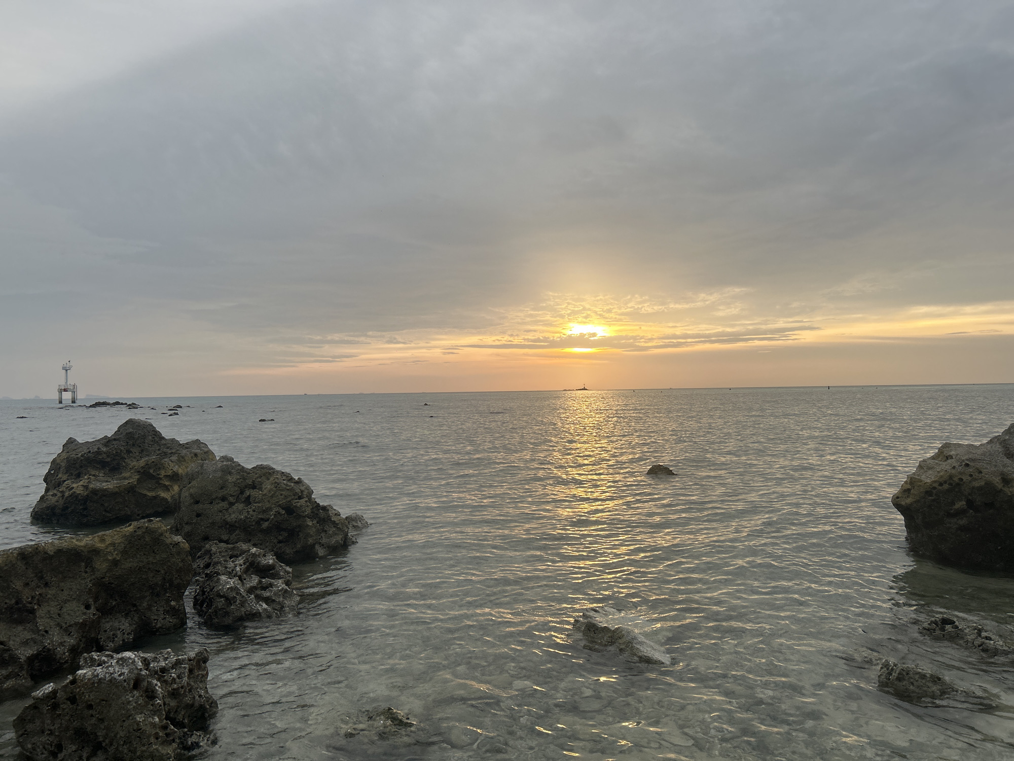 This is a photograph of a serene beach at sunset on Ko Pha-ngan District, Thailand. The sky is overcast with scattered clouds, and the sun is low on the horizon, casting a warm, golden glow across the sea. The water is calm and shallow, revealing a sandy bottom with scattered rocks and small waves gently lapping at the shore. In the foreground, dark, rough-textured rocks are partially submerged in the water, adding a rugged element to the scene. In the distance, a small lighthouse stands on a rocky outcrop, and a distant boat is visible on the horizon. The overall impression is one of peace and tranquility, with the natural beauty of the seascape bathed in the soft light of the setting sun.