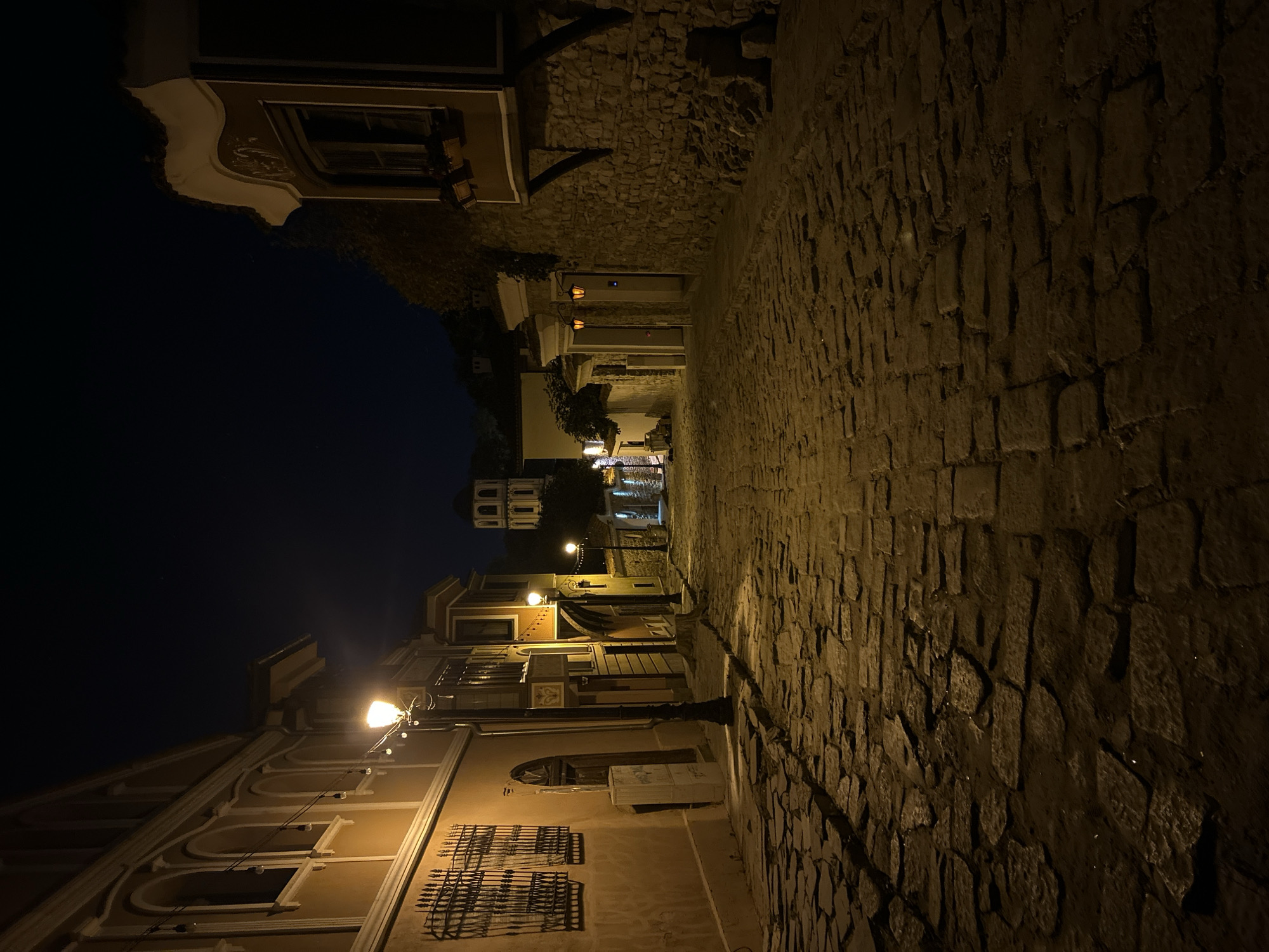 The image depicts a narrow, cobblestone street at night in the Old Town of Plovdiv, Bulgaria. The street slopes gently upwards and appears to be lined with historic buildings on both sides. Several street lamps cast a warm glow on the cobblestones, creating a dramatic play of light and shadow.
On the left side of the street, a building with a yellow facade is visible. It features arched windows with ornate metal grilles. A street lamp stands prominently in front of this building. Further up the street on the left, more buildings with yellow and ochre facades are visible, some with decorative architectural details.
On the right side of the street, a building with a stone facade is prominent. It has a distinctive overhanging upper story with a window box. Further up, another building with a light-colored facade and a stone base is visible, featuring two illuminated lantern-style lights at an entrance.
In the distance, at the top of the incline, a white tower with an arched design is visible against the dark night sky, suggesting a church or other historical monument. The overall atmosphere is quiet and somewhat mysterious, evoking a sense of historical charm and tranquility. There are no people or animals visible in the image. The environment is urban, with a focus on historical architecture and street elements.