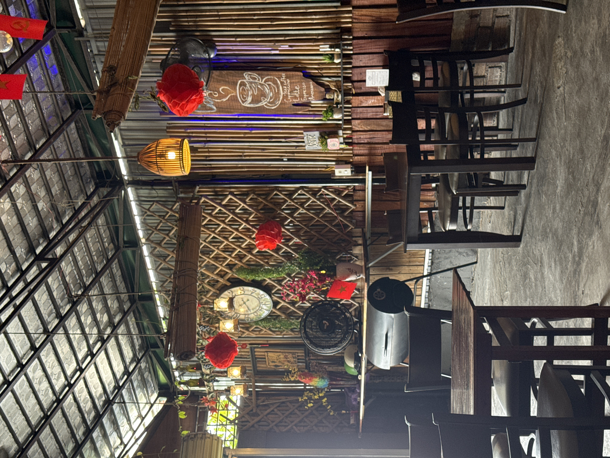 The image depicts the interior of a cafe, likely in Nha Trang, Vietnam, characterized by a rustic and natural aesthetic. The ceiling is constructed with corrugated metal panels supported by dark metal beams, and a bamboo-like blind is suspended from the structure. The walls are predominantly adorned with bamboo poles arranged vertically and diagonally, creating a textured backdrop.
The space is furnished with several wooden tables and chairs, suggesting a dining or seating area. One table in the foreground is made of dark wood and has four chairs around it, with two of them clearly visible, each with a dark leather seat and back. Another table is visible in the background, also dark wood, with chairs around it.
Decorative elements include red paper lanterns hanging from the ceiling, a vintage-style wall clock, a black electric fan, vases with flowers, and a framed picture on the bamboo wall. A prominent sign on the right side of the wall features a hand-drawn coffee cup with text listing coffee types like "Iced Coffee," "Cappuccino," "Latte," "Espresso," and "Americano." Other small decorative items, such as plants in pots and what appears to be a miniature religious figurine, are also present on shelves. A large, dark grill or smoker is situated against the bamboo wall, partially visible in the mid-ground.
The lighting appears to be a mix of ambient light filtering from outside and from artificial sources like the hanging lamp and the lanterns. The overall impression is of a relaxed, casual atmosphere, typical of a cafe in a warm climate, with a strong emphasis on natural materials and a touch of cultural decoration. There are no people visible in the image.