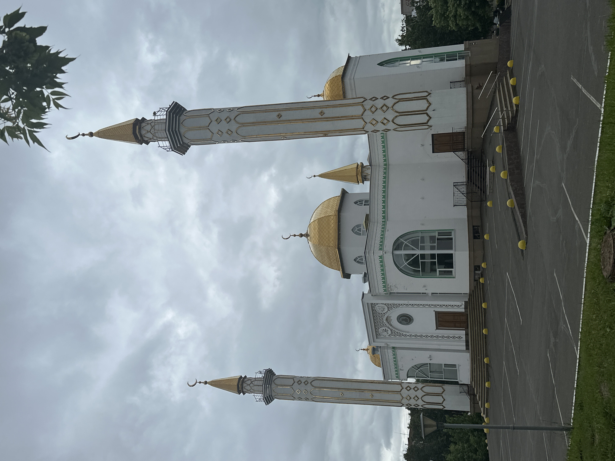 This forensic description details the Izhevsk Central Mosque, located in Izhevsk, Russia, under an overcast sky. The prominent white architectural structure features a central golden dome, two smaller golden domes, and two tall, slender minarets, all topped with golden spires and crescent moon finials. The minarets exhibit vertical segments with repeating golden diamond-shaped patterns. The mosque's facade includes large arched windows, some with green frames, and decorative elements above the main entrance featuring intricate white and gold Islamic geometric and calligraphic designs.
The building is situated on an elevated platform, accessed by multiple sets of stairs with dark handrails, leading to various entrances, including a double wooden door at the main facade. The foreground consists of an empty asphalt parking lot with white painted lines delineating parking spaces. Numerous spherical yellow bollards are arranged along the perimeter of the mosque's elevated base and within the parking area. A gray street lamp stands on the left side of the parking lot. A strip of green grass is visible in the immediate foreground, with scattered trees and foliage forming a backdrop around the mosque and in the distance. The sky is uniformly gray and heavily clouded. No individuals are present in the scene.