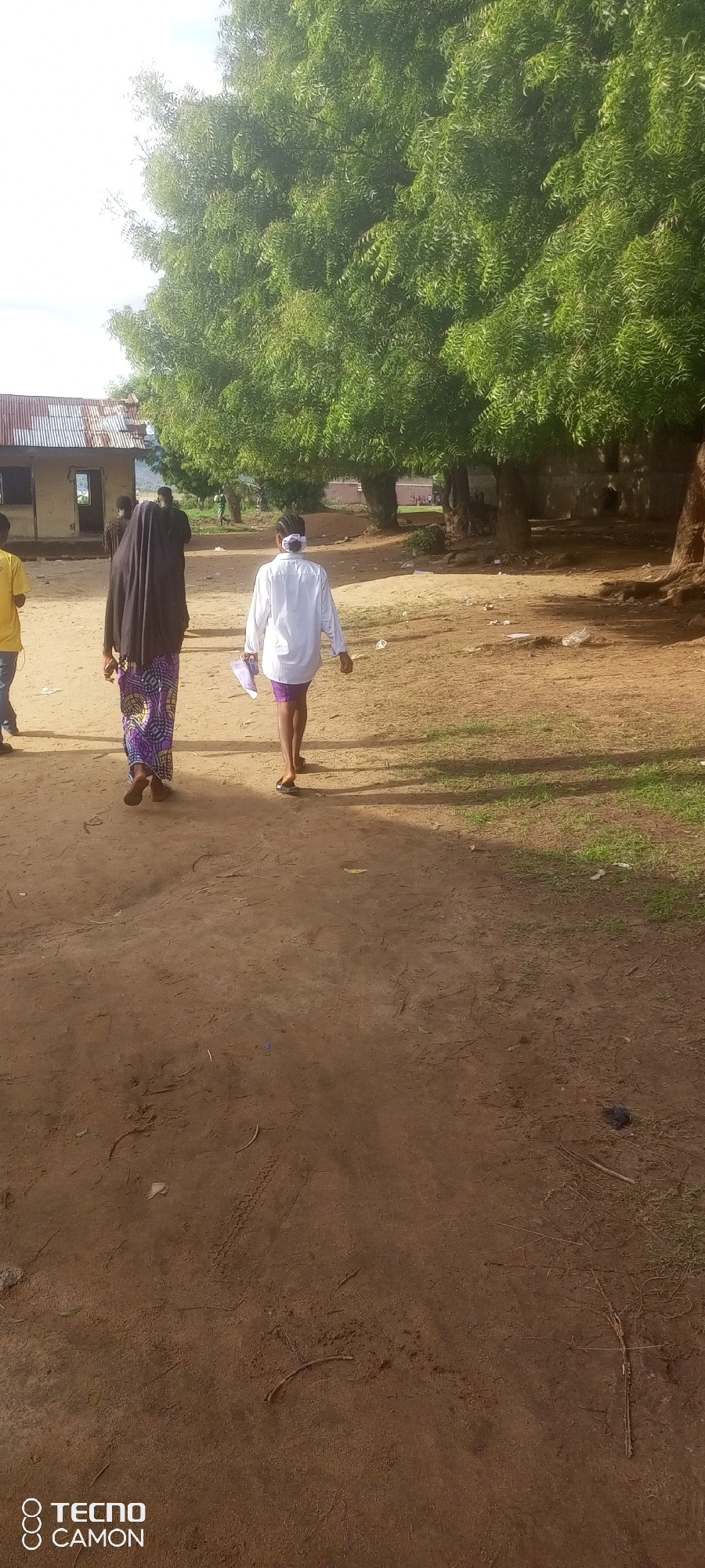 The image depicts an outdoor scene in Jalingo, Nigeria, with a dirt path leading through a semi-arid environment characterized by sparse vegetation and trees with dense green foliage. In the foreground, two individuals are walking away from the camera along the path. The person on the left wears a long, dark head covering and a patterned, floor-length skirt, while the person on the right wears a white long-sleeved shirt and purple shorts. Both are barefoot or wearing dark open-toed footwear. To the left of the path, a simple, weathered building with a corrugated metal roof is visible, with another person in a yellow shirt standing near it. Further in the background, more trees and a few other people are discernible, suggesting a communal or residential area. The ground is dry and dusty, with scattered debris including leaves and what appear to be pieces of paper or plastic. The lighting indicates it is daytime, with sunlight casting shadows from the trees. The overall impression is one of a rural or semi-urban setting in Nigeria during the day.