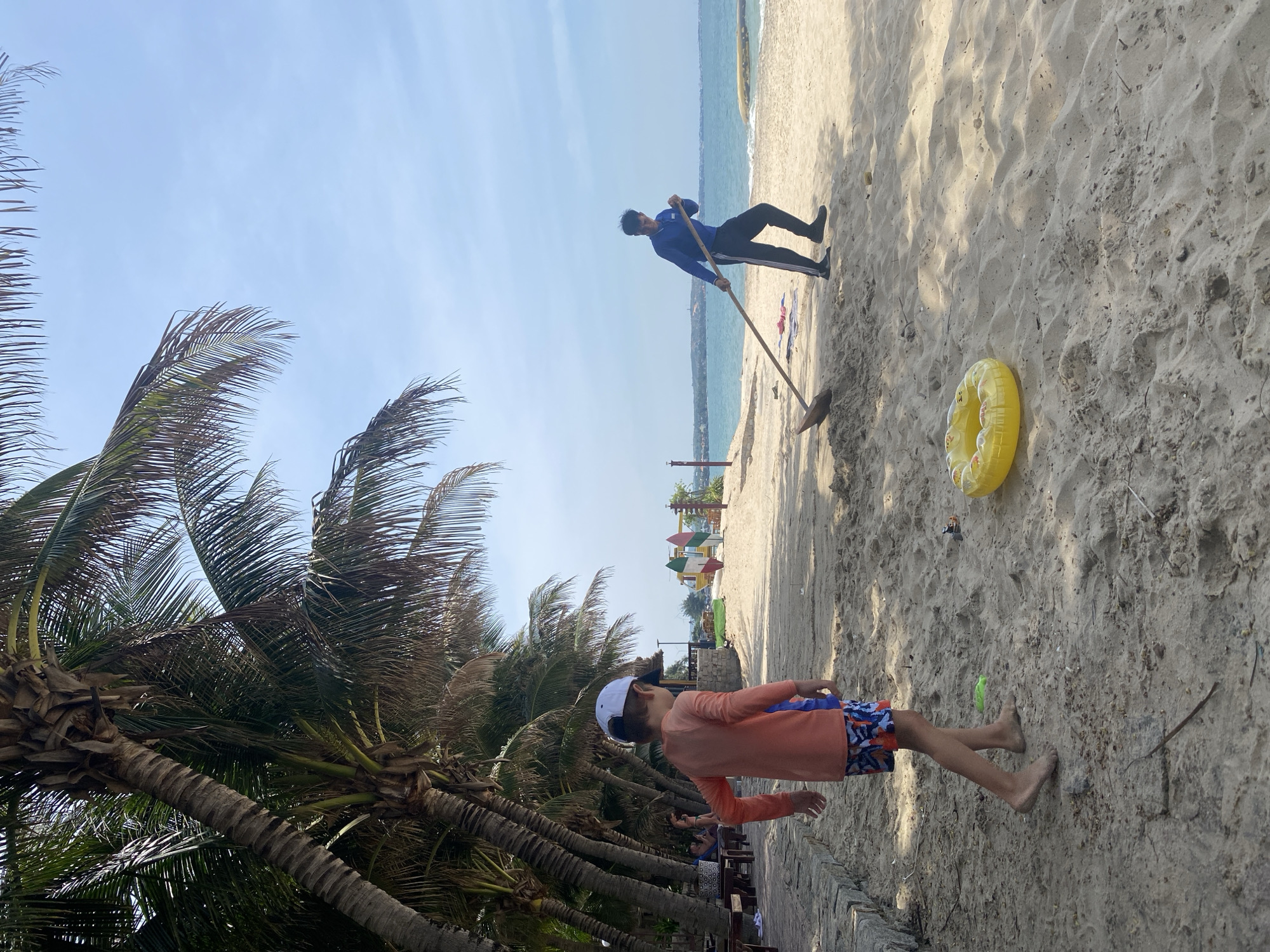 The image depicts a sandy beach scene under a clear blue sky, with palm trees lining the left side of the frame. In the foreground, a young boy wearing an orange long-sleeved shirt, patterned shorts, and a white baseball cap stands facing away from the camera, his left foot slightly raised as if walking. To the right, a man in a blue long-sleeved shirt and dark pants is using a shovel to move sand. A yellow inflatable ring lies on the sand in the middle ground between the two figures. The ocean is visible in the background. The location is identified as Phu Thuy Ward, Vietnam.