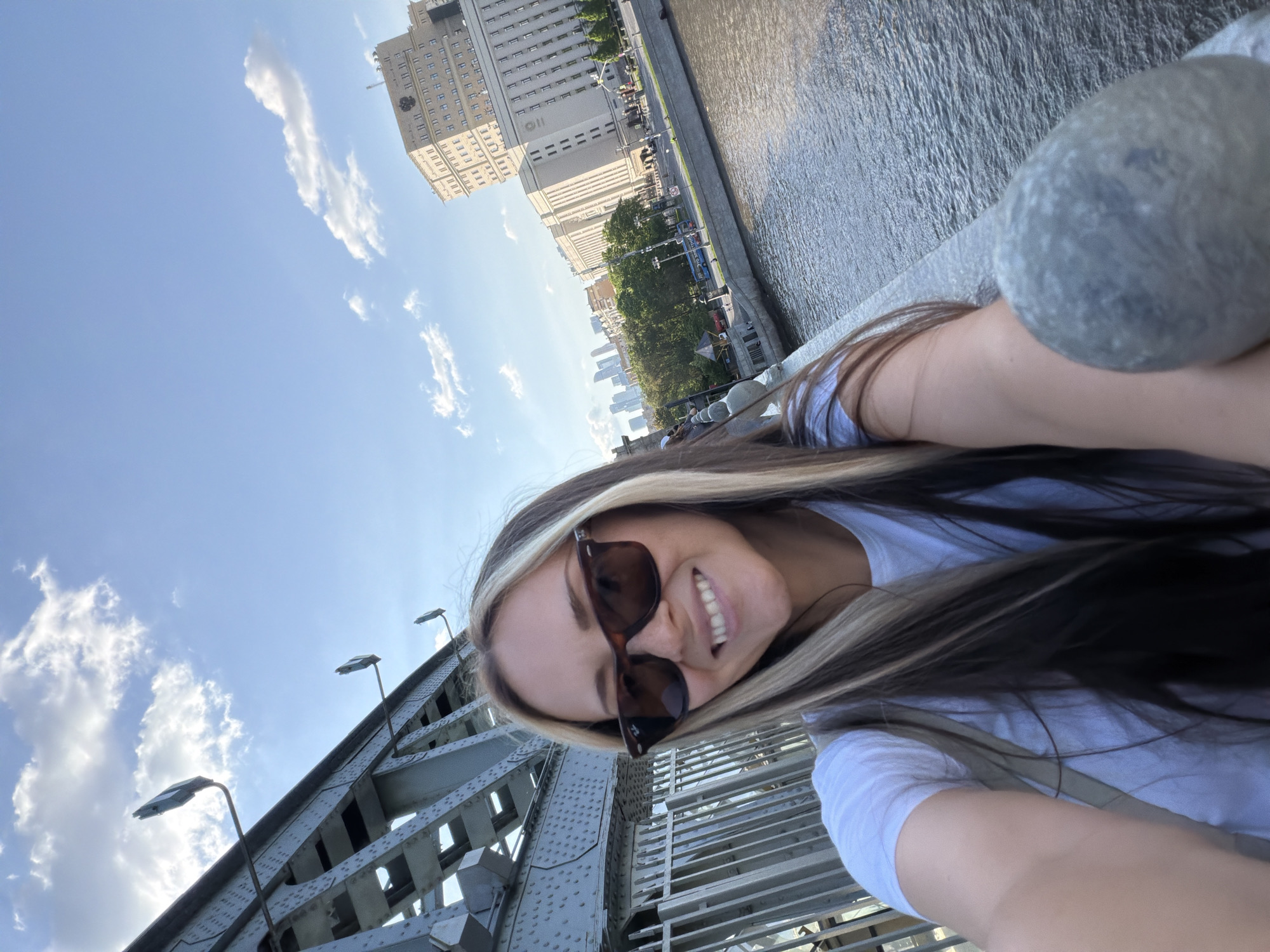 A young woman is taking a selfie while standing on a bridge in Moscow, Russia. In the background, a large building with a distinctive facade can be seen, identifiable as one of the Stalinist skyscrapers, potentially the Ministry of Foreign Affairs. The bridge has a metal structure with rivets and includes streetlights. To the side of the bridge is a river with a metal railing. The sky is blue with some clouds. The woman is wearing sunglasses and a white shirt, and has long hair with blonde highlights. She is smiling at the camera. A gray, amorphous object is present at the upper right edge of the frame.