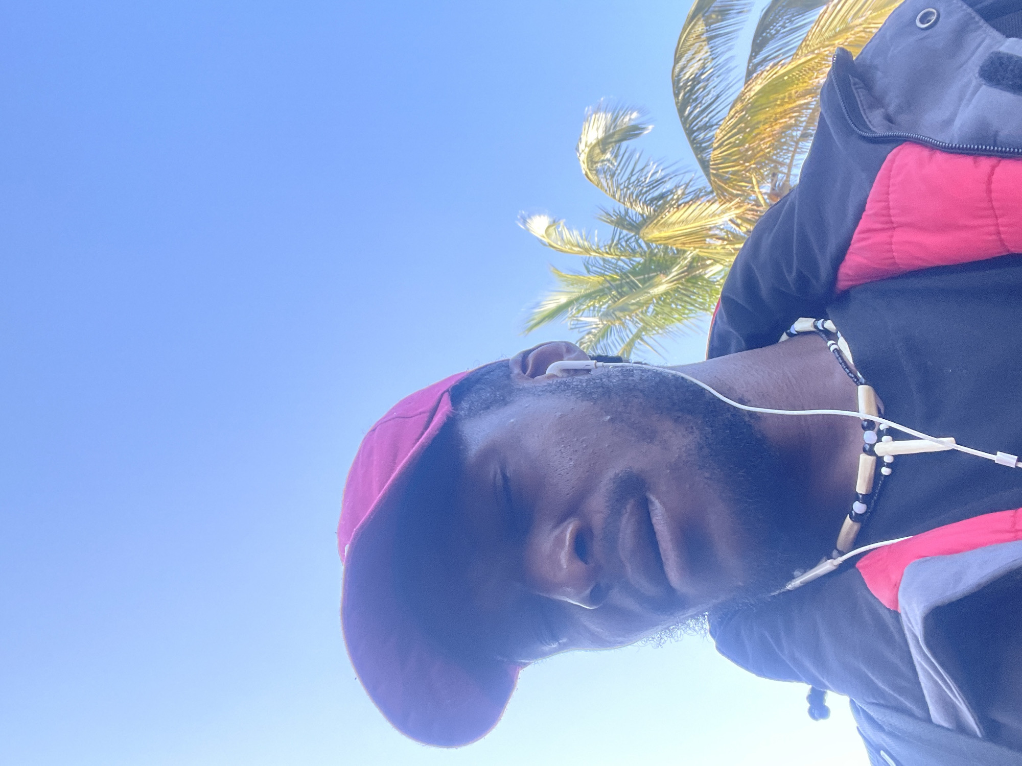 The image is a selfie taken outdoors on a bright, clear day, likely in Jos, Nigeria, indicated by the presence of palm trees and clear blue skies, which are common in the region. The subject is a dark-skinned man wearing a red baseball cap, a black t-shirt, and a jacket with a blue exterior and a red lining. He is also wearing white earphones and a beaded necklace. The man's eyes are closed, and he appears to be looking down. In the background, several palm trees are visible against the bright blue sky. The overall lighting suggests it is daytime, with the sun casting a slight glare on the subject's face.
