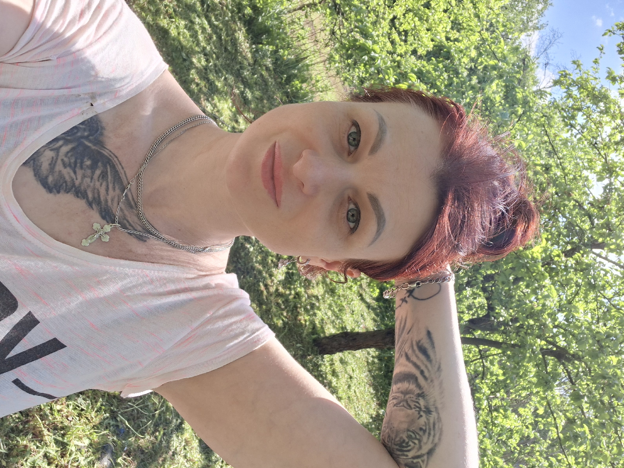 A woman with red hair and green eyes is in an outdoor setting. She has a tattoo on her upper chest, and a tattoo of a tiger on her right arm. She is wearing a light pink shirt and a silver chain with a cross pendant. The background includes green trees and grass, suggesting a park or natural area. The location is Сваткаўскі сельскі Савет, Belarus.