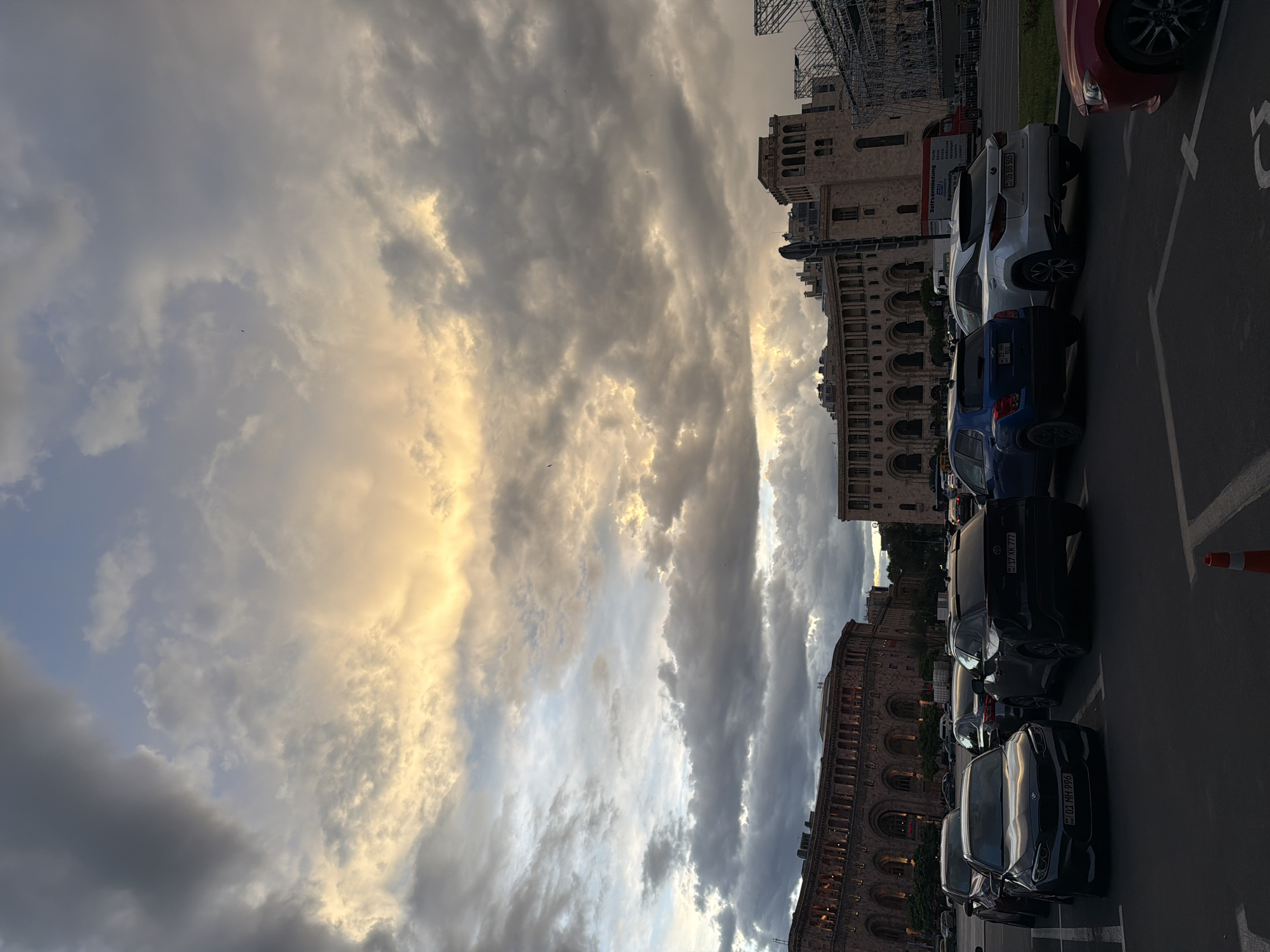 The image captures a street scene in Yerevan, Armenia, showcasing a row of parked cars alongside buildings. The sky overhead is filled with clouds, exhibiting variations in color and brightness. The buildings display architectural features, with visible windows, arches, and detailing. The cars vary in color and model. The street appears paved and lined with parking markers. The overall impression is of a city landscape under an overcast sky.