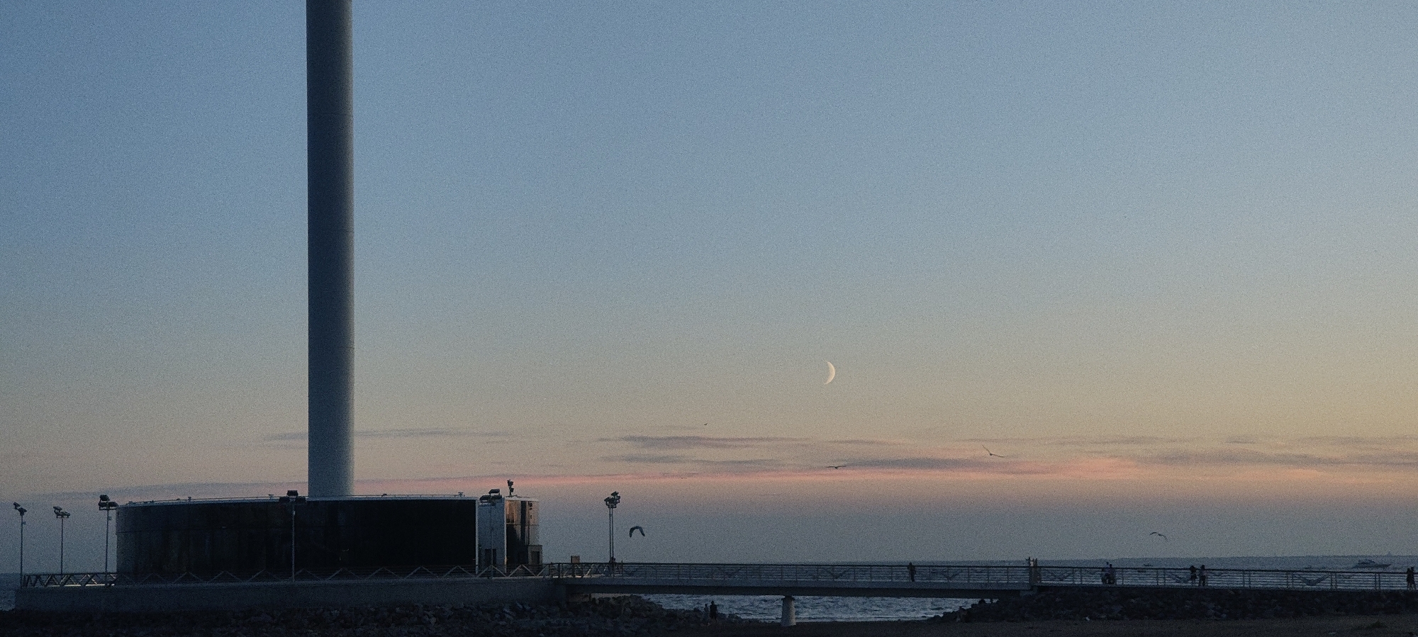 A wide shot captures a pale crescent moon in a light blue sky with a gradient of soft pink and orange hues near the horizon. On the left, a tall, cylindrical white structure stands prominently, its base a dark, circular building with a walkway extending to the right. This walkway, with railings and scattered figures, stretches over the water. In the foreground, a dark, rocky shore is visible. The scene appears to be a coastal city location at dusk or dawn, with a serene atmosphere and a sense of calm.