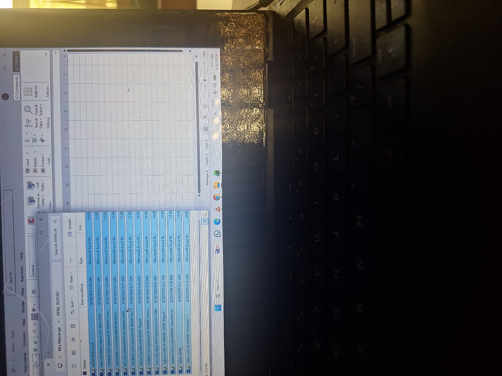 The image depicts a close-up view of a laptop screen displaying a Microsoft Excel spreadsheet. The spreadsheet is titled "APRIL REPORT" and is located within a folder named "Mrs Madanga." Several files are listed, each with a name, date modified, type, and size. The files appear to be Microsoft Excel documents. The date modified for most of the files is June 30, 2026. The screen is showing a portion of the spreadsheet and the Excel ribbon interface, which includes options for "File," "Formulas," "Data," "Review," "View," "Automate," and "Help." Below the ribbon, there are options for "Page Layout" and "General." The laptop keyboard is visible at the bottom of the image, with some keys illuminated by ambient light. The screen itself has some visible damage or residue on the bottom edge, appearing as a textured, dark material. The overall scene suggests a work or study environment. The location of Jalingo, Nigeria is not directly represented in the image content.
