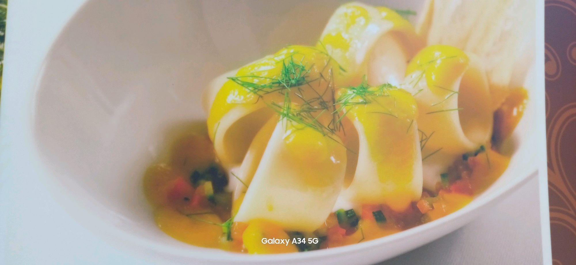 A close-up, slightly blurred, overhead shot features a white ceramic bowl filled with a culinary dish. The dish consists of several pieces of what appear to be white, rolled or folded food, possibly pasta or seafood, coated in a vibrant yellow sauce. Finely chopped green herbs, likely dill, are sprinkled over the top, adding a delicate garnish. The sauce has a glossy sheen, and visible throughout the dish are small, colorful diced vegetables, including hints of red and green. The background is a soft, warm light, indicating an indoor setting. The bottom right corner of the image displays the text "Galaxy A34 5G," indicating the device used for capture. This image is not associated with Abano Terme, Italy.