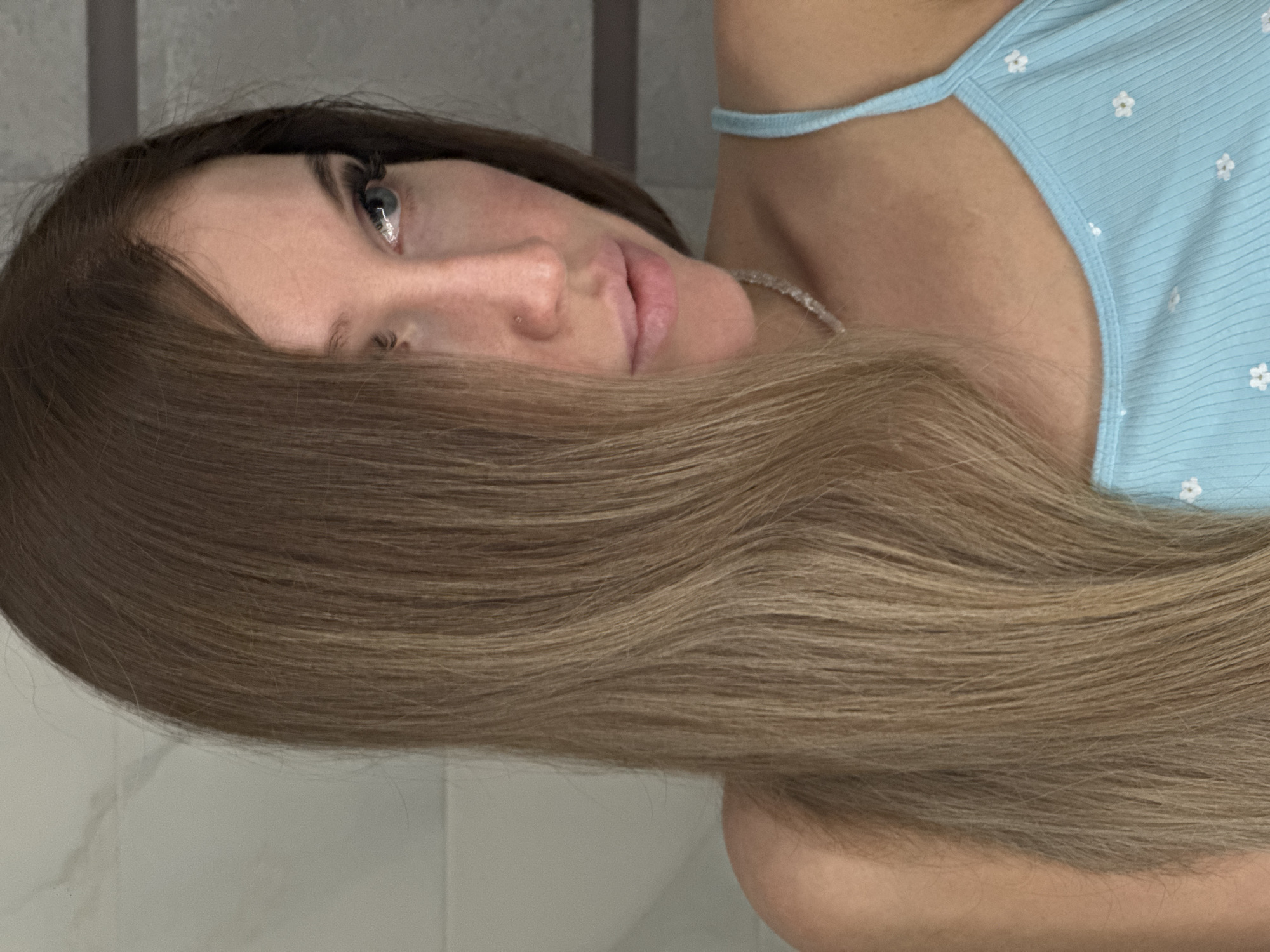 A close-up shot focuses on a woman's face and upper torso, with her head turned to the right, looking away from the camera. She has long, brown hair that falls over the left side of her face, obscuring part of her eye. Her skin is fair, and she is wearing subtle makeup, including mascara and what appears to be a light foundation. A small nose stud is visible. Her lips are slightly parted, and she has a delicate necklace around her neck. She is wearing a light blue, ribbed tank top with a subtle floral pattern. The background consists of a blurred, tiled wall and a darker section on the right, possibly a cabinet or door. There are no discernible elements that would allow for pinpointing a specific location within the city of Chaikovsky, Russia.
