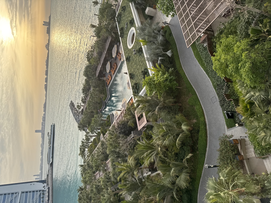 The image displays an elevated view of a coastal resort landscape in Dubai, United Arab Emirates, with the distinctive Burj Al Arab Jumeirah hotel visible on the far left. The scene is illuminated by the golden light of the sun, positioned low in the sky, creating a bright reflection across the surface of the Arabian Gulf to the right.
In the foreground and midground, a densely landscaped area features numerous palm trees of various species, tropical plants, and green ground cover. A winding, paved pathway constructed from grey interlocking pavers traverses this green space. Several structures with wooden slatted pergolas or roofs are integrated into the landscaping. A large, irregularly shaped swimming pool with light blue water is visible, surrounded by approximately a dozen orange sun loungers and several white parasols. Adjacent to the pool area, a flat green roof with a distinct circular opening is present. One individual is observed walking on the paved pathway in the middle-right section of the image, moving away from the viewer.
The background features the turquoise waters of the Arabian Gulf. A man-made rock breakwater extends from the shoreline into the sea. A long, white vessel is docked in the water near the base of the Burj Al Arab. The distant horizon across the gulf reveals a sprawling cityscape with multiple high-rise buildings and land formations, including elements consistent with the Palm Jumeirah.