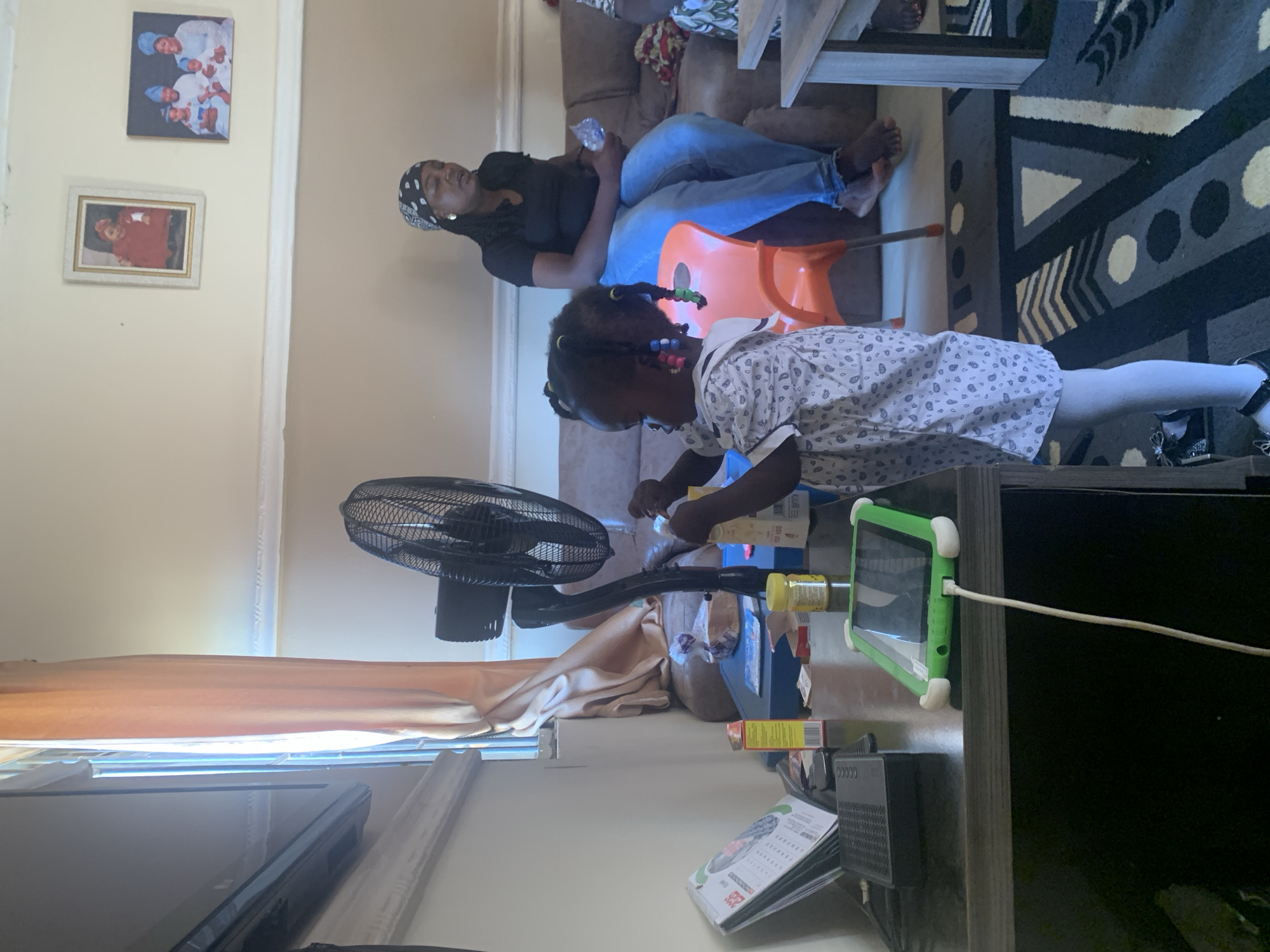 This image depicts an indoor scene, likely a living room, in the city of Gura Top, Nigeria. In the foreground, a young Black child stands in front of a black desk, interacting with a fan and a tablet. The child is wearing a light-colored patterned dress and white tights. To the right, a Black woman with braided hair and a headscarf is seated on a brown couch. She is wearing a black t-shirt and blue jeans. The environment includes a desk with various items, including a tablet, a fan, a calendar, and what appear to be food items or containers. The background shows a couch, a window with curtains, and framed pictures on the wall, one of which shows a family. The lighting suggests it is daytime.