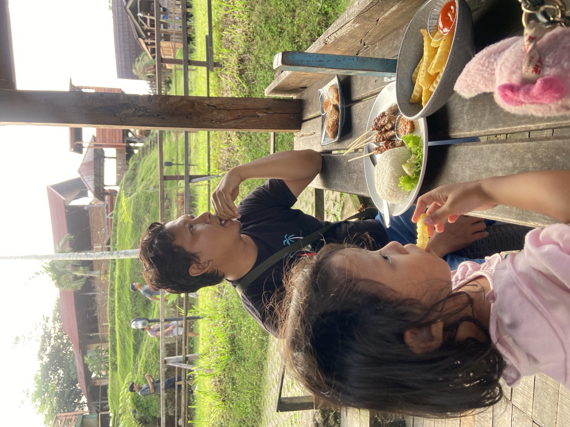 The image shows a man and a young child seated at a wooden table outside, possibly at a restaurant or eatery, in Selotapak, Indonesia. The man is eating something with his hand. The child is eating a french fry. There are plates of food on the table, including what appears to be satay with rice and lettuce, and a container of french fries with ketchup. The background features buildings, greenery, and a walkway or bridge. Other people can be seen walking in the background.