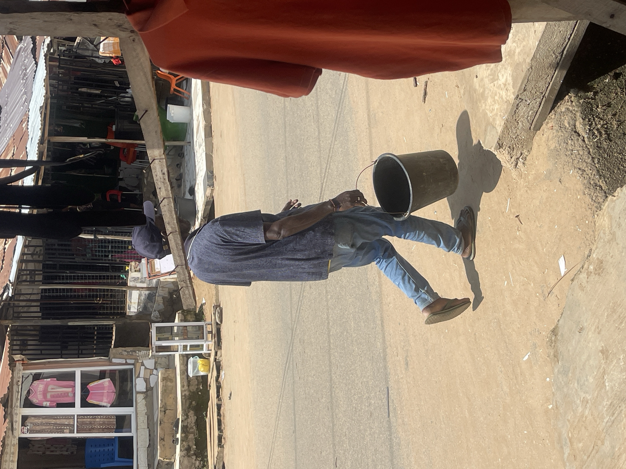 A man wearing a dark blue short-sleeved shirt, blue jeans, a dark blue cap, and brown sandals walks along a dirt road in Abuja, Nigeria. He carries a dark gray bucket in his right hand. The man is walking from left to right in the frame. In the background, there are market stalls with various goods displayed, including clothing. A blue chair is visible outside one of the stalls. The ground is a mix of dirt and paved surfaces. The scene appears to be during daylight hours, with clear shadows cast by the man and the structures.
