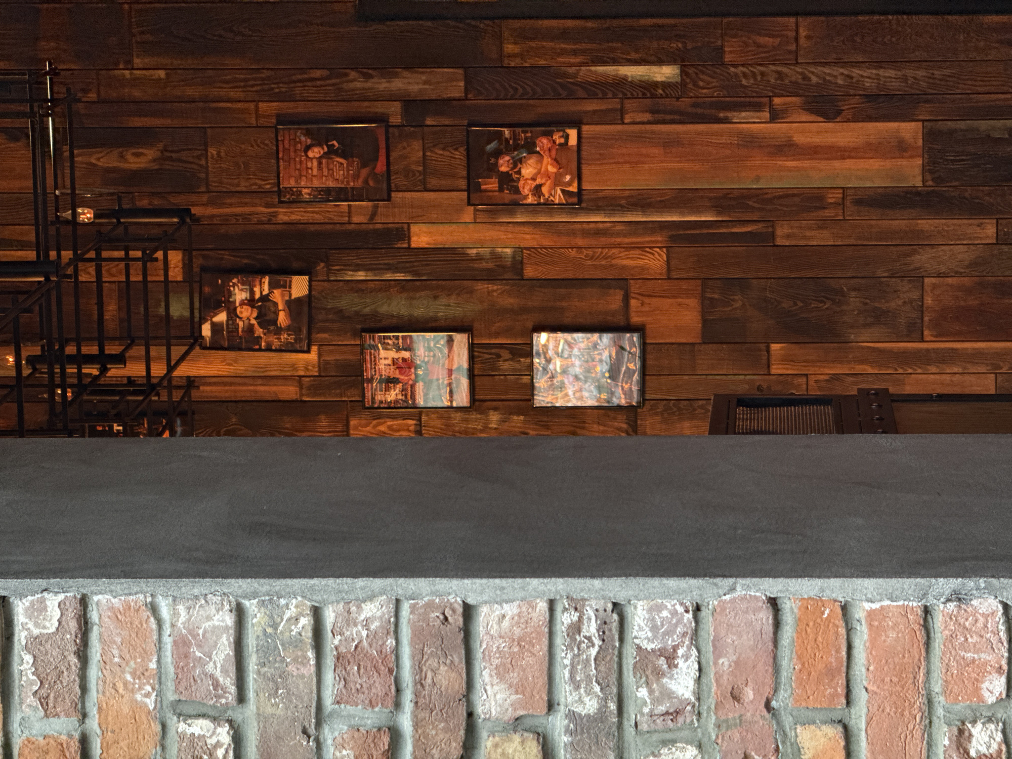 The scene depicts an interior commercial establishment in Izhevsk, Russia, characterized by a tripartite vertical composition. The leftmost section features an exposed brick wall with reddish-orange bricks and visible white mortar, exhibiting some surface discoloration. Adjacent to this, a smooth, dark grey vertical pillar or wall segment stands centrally. The rightmost and dominant section is a paneled wall constructed from varying widths and lengths of dark, warm-toned wooden planks, displaying prominent grain patterns.
An industrial-style lighting fixture, composed of dark metal bars forming an open cage structure, is suspended from an unseen ceiling at the top right, containing multiple illuminated, warm-glowing bulbs. Five framed rectangular images are mounted on the wooden wall. The upper left frame displays a portrait of a woman with dark hair, wearing a dark apron, with her arms crossed. The upper right frame contains a portrait of a woman with dark hair, dressed in dark attire, seated on a red surface, against a background featuring a brick wall. The middle-left and lower-left frames each contain abstract or impressionistic artworks with various colors. The lower-center frame presents a portrait of a bearded man wearing an apron, holding raw meat or food items. The overall environment suggests a culinary or food service context.