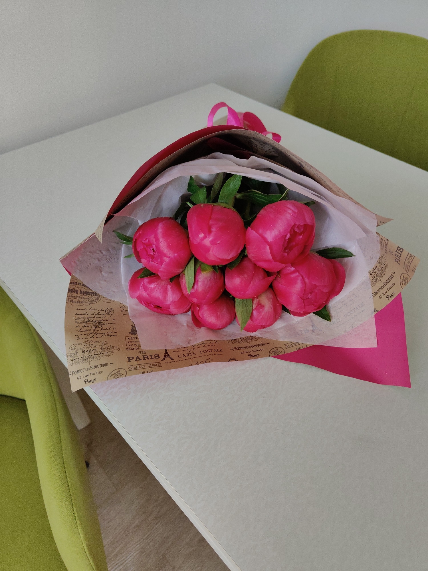 A vibrant bouquet of pink peonies rests on a light-colored table surface in an indoor setting. The peonies, mostly in bud form, display rich fuchsia hues with visible green leaves and small water droplets on some petals, suggesting freshness.
The flowers are meticulously wrapped in layers. An inner layer of soft white tissue paper cradles the blooms, encased by a larger sheet of brown kraft paper featuring French-themed text and vintage designs, reminiscent of an old postcard or newspaper. Prominent phrases include "PARIS A CARTE POSTALE," "FABRIQUE DE BIJOUTERIE 62 Rue Turbigo PARIS," and "CATALOGUE ILLUSTRE," along with "TOULOUSE" and "ROVE ROUST," establishing a distinct Parisian aesthetic. A bright pink paper layer is visible beneath the brown wrapping, and a matching pink ribbon is tied at the top of the bouquet.
In the background, the soft, curved backs of two lime green chairs are partially visible, one on the right and one on the bottom left, suggesting a comfortable domestic or office environment. The lighting is bright and even, indicating daytime. The scene evokes a sense of thoughtful gifting, celebration, or home decor. No people are visible in the image.