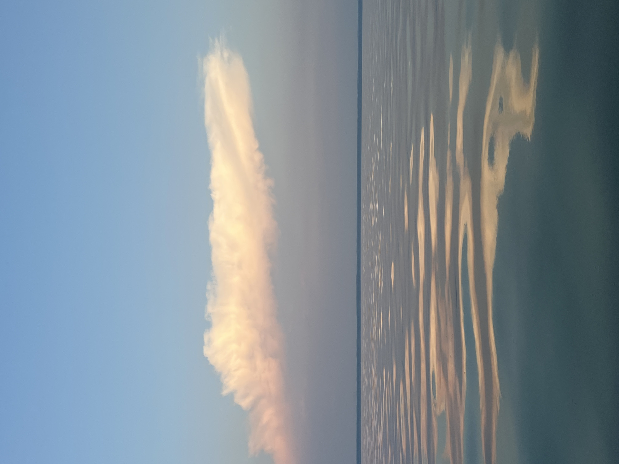 The image depicts a serene natural landscape at what appears to be either dawn or dusk. The sky is a soft gradient of blue, transitioning to a pale pinkish hue near the horizon, where a single, elongated cloud is illuminated with warm, peachy tones. Below the sky, a thin, dark line suggests a distant landmass or the horizon itself. The foreground is dominated by a calm body of water, likely a lake, with gentle ripples reflecting the colors of the sky and cloud. These reflections create abstract patterns of light and shadow on the water's surface, ranging from pale pinks and oranges to muted blues and greens. There are no people, man-made structures, or specific landmarks visible that would pinpoint a location within Borovoye, Russia. The scene is a minimalist representation of natural beauty, focusing on the interplay of light, sky, and water.
