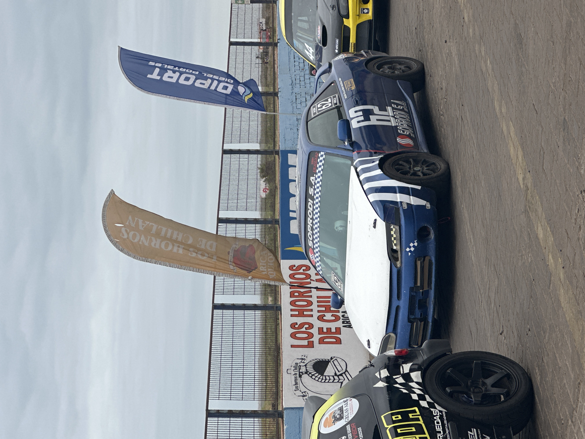 The image shows a section of a racetrack in Arica, Chile, where several racing cars are positioned on the asphalt surface. In the background, behind a metal fence, is a sign with the words "LOS HORNOS DE CHILLAN ARICA" in large red letters, with smaller text below and a logo of "Los hornos de Chillán". Flags with the words "DIPORT DIESEL PORTALES" (blue) and "LOS HORNOS DE CHILLAN ARICA" (beige) are also visible. The racing cars are in various colors with racing numbers. Car details include racing sponsorships such as "SSSOPRODI S.A." and "RUEDAS EGO PORTALES #2428". A partial view of the sky suggests it is a cloudy day.