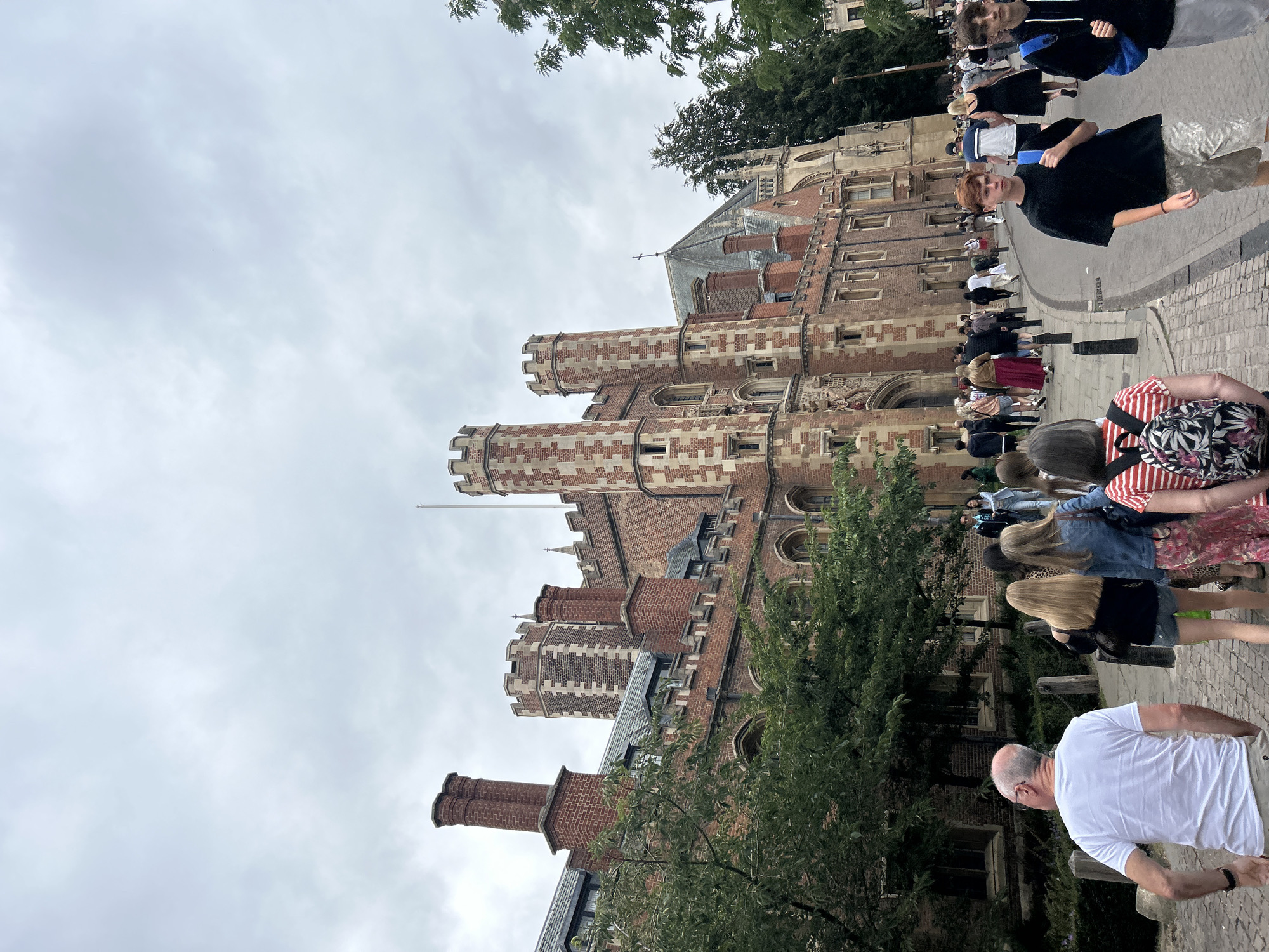 The image depicts a street scene in Cambridge, United Kingdom, featuring the imposing architecture of what appears to be a historic college building, likely part of the University of Cambridge. The building is constructed of red brick with distinctive white stone detailing around the windows and parapets, and features prominent turrets. A group of people are walking along a cobblestone path in front of the building, some in the foreground and others further down the street. The sky is overcast, suggesting a cloudy day. Green trees are visible on the left side of the image, bordering the path and the building. The people in the image are varied in age and attire, some appearing to be tourists or students.