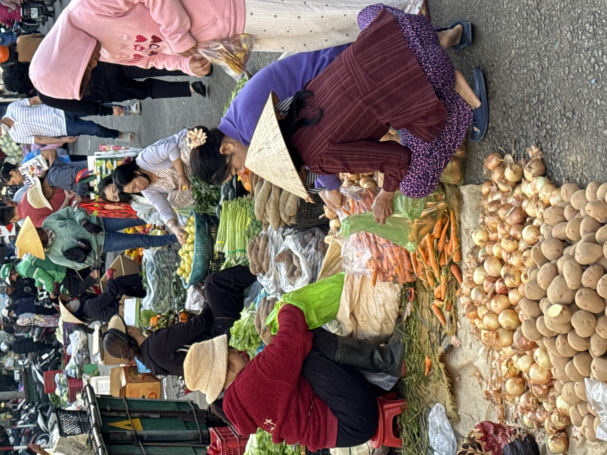 The image depicts an outdoor market scene in Da Lat, Vietnam, likely a produce market. Several individuals, predominantly women, are engaged in activities related to selling and purchasing vegetables. One woman in a red sweater and a bucket hat is holding a green plastic bag, while another wearing a purple top and a conical hat is interacting with produce. A woman in a pink hoodie is also present, observing or participating in the market activities. The foreground displays a variety of vegetables, including potatoes, onions, and carrots, arranged on the ground. In the background, there are glimpses of other market stalls, motorcycles, and a crowd of people, suggesting a bustling environment. The ground appears to be asphalt or a similar paved surface, common in urban settings. The overall impression is one of a lively, everyday commercial exchange of goods.