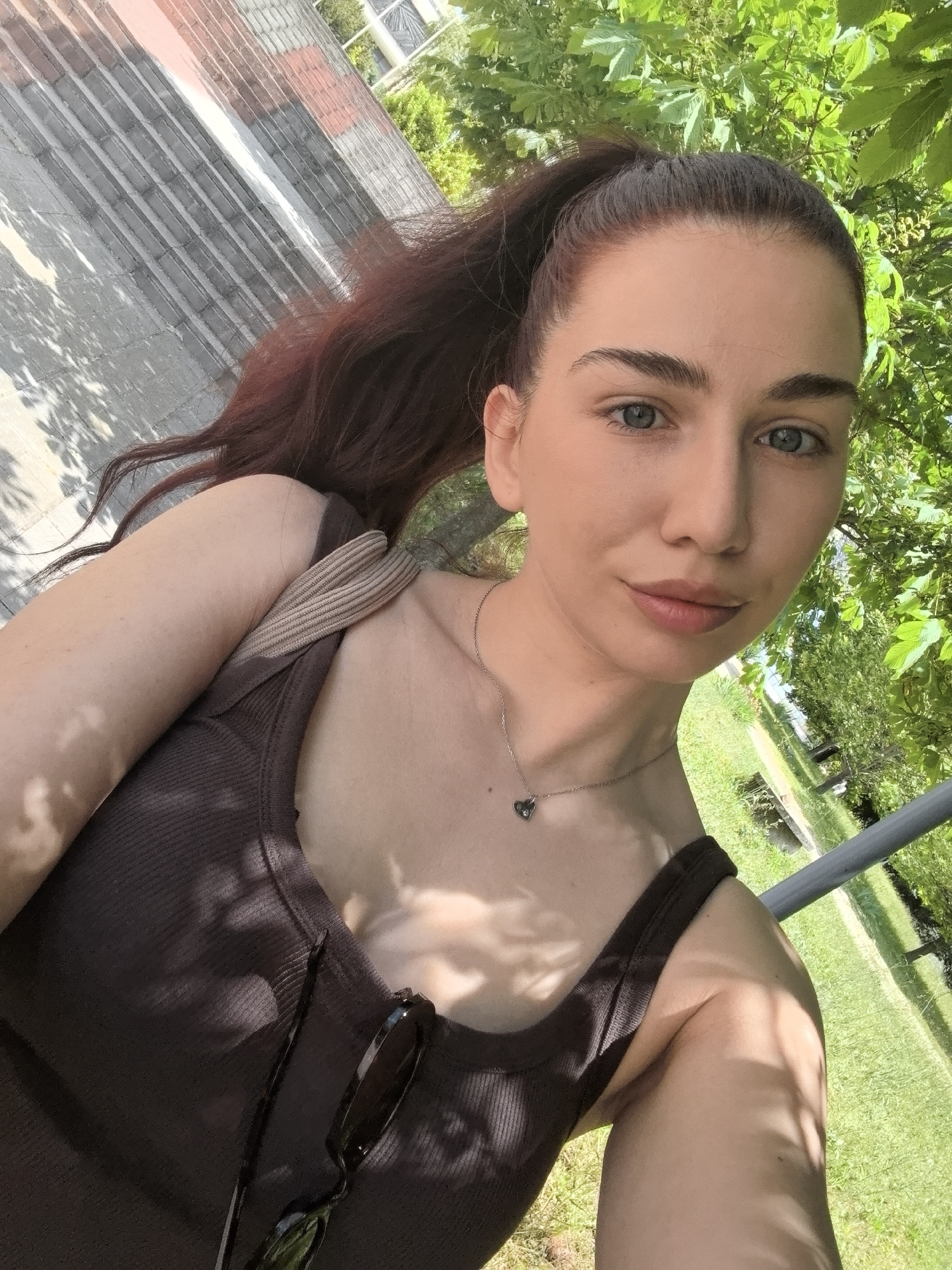 A selfie of a young woman with auburn hair pulled back into a high ponytail, wearing a brown tank top and a silver necklace with a heart pendant. Sunglasses are hooked on her tank top. She is outdoors, with dappled sunlight from trees overhead creating shadows on her skin and clothing. In the background, there is a building with a brick and concrete facade and green grass. The location is in an urban setting, specifically the city of Unknown, Unknown.