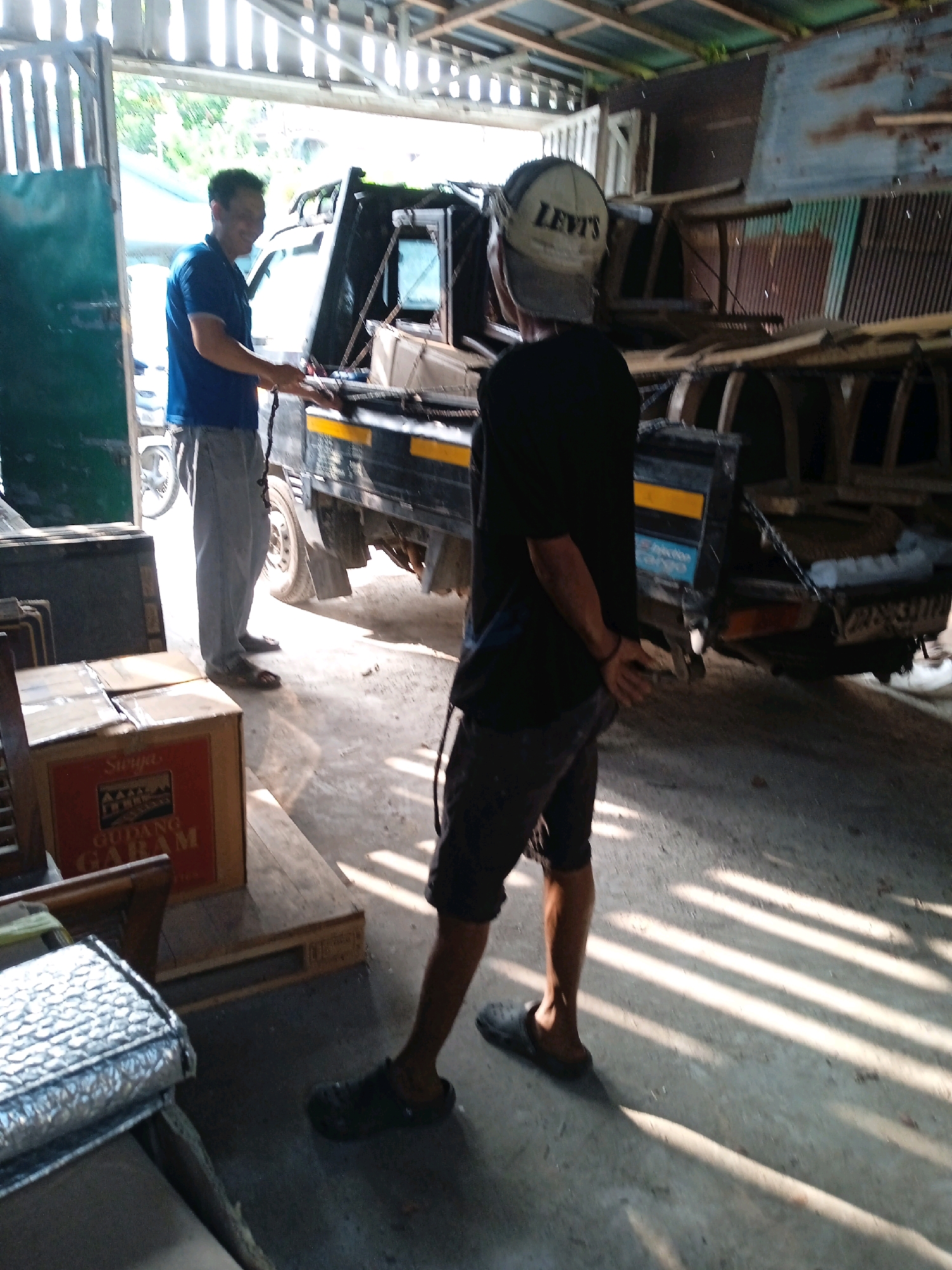A man in a blue t-shirt and light grey pants stands in what appears to be an open-air workshop or garage in Banjarmasin, Indonesia. He is facing a black truck with a yellow stripe on its side. Another person, wearing a black t-shirt, dark shorts, and a baseball cap, stands with their back to the camera, looking at the truck. The environment is cluttered with various objects, including wooden pallets, metal sheets, and what looks like salvaged parts. Sunlight streams in from the open entrance, casting strong shadows on the concrete floor. A large cardboard box with red lettering is visible on a wooden pallet in the foreground.