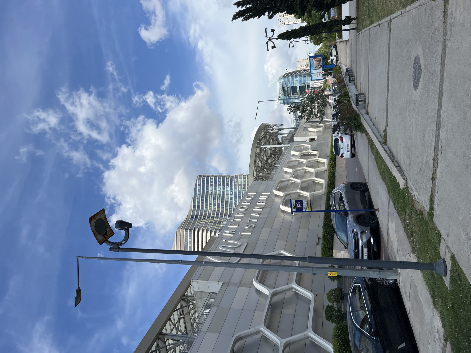 The image captures a daytime, eye-level, exterior view of the Metropol Istanbul shopping mall and office complex in Ataşehir, Turkey. The foreground features a concrete sidewalk with a grassy verge on the left, bordered by a low concrete retaining wall. A street lamp post with a damaged solar panel is prominently positioned on the sidewalk. To the left of the sidewalk, a black sedan is parked, and a silver SUV is parked behind it. Further down the sidewalk, there are several concrete benches.
The main subject, the Metropol Istanbul building, dominates the midground. It's a large, modern structure with a silver facade featuring a hexagonal pattern. Illuminated white text on the building reads "METROPOL ISTANBUL". To the right of the main building, a tall, curved skyscraper with multiple floors of windows rises into the sky.
In the background, more modern buildings are visible, including one with "ACIBADEM" signage. The sky is a bright blue with scattered white clouds. The overall scene depicts an urban environment with architectural elements and clear weather.