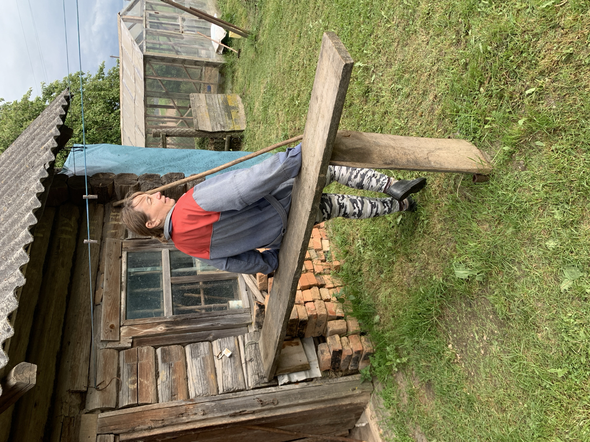 This image depicts a rural scene in Відзаўскі сельскі Савет, Belarus, featuring a weathered wooden house with a person seated on a bench outside.
In the foreground and midground, a woman is seated on a rough-hewn wooden bench that extends from the left side of the frame towards the right. She is oriented with her back partially to the viewer, looking towards the left side of the image (viewer's right). Her hair is light brown and pulled back, and she wears a two-toned jacket, red across the shoulders and upper back, transitioning to gray on the sleeves and lower torso. Her pants are camouflage pattern in shades of gray and white. She wears dark shoes and her left foot is visible. Her right arm is bent at the elbow, resting on the bench, and her left hand is visible, appearing to hold a long, slender wooden stick that leans against the blue tarp/fabric.
The bench itself is a thick, unpolished wooden plank, supported by a vertical wooden post on its right side, anchored in the grass. Underneath the left end of the bench, near the house, is a stack of reddish-brown bricks.
To the left of the woman and bench is the exterior of an old, rustic wooden house constructed from horizontal logs. The house features a window with multiple panes, visible behind the woman's left shoulder. Above the window, a light blue clothesline with a white clothespin is stretched horizontally across the wooden wall. A blue tarp or fabric hangs vertically from the eaves of the house, partially obscuring the space between the house and the background. The roof of the house is made of corrugated gray material, possibly asbestos cement, with a weathered appearance.
In the background, behind the house and to the right, two transparent greenhouses with wooden frames are visible. The one on the right is larger and clearer. Beyond the greenhouses and house, green trees are visible, and the sky is overcast, suggesting a cloudy day. The ground in the foreground and midground is covered in green grass with some bare patches of soil. A thin metal pipe protrudes from the grass near the right support of the bench.