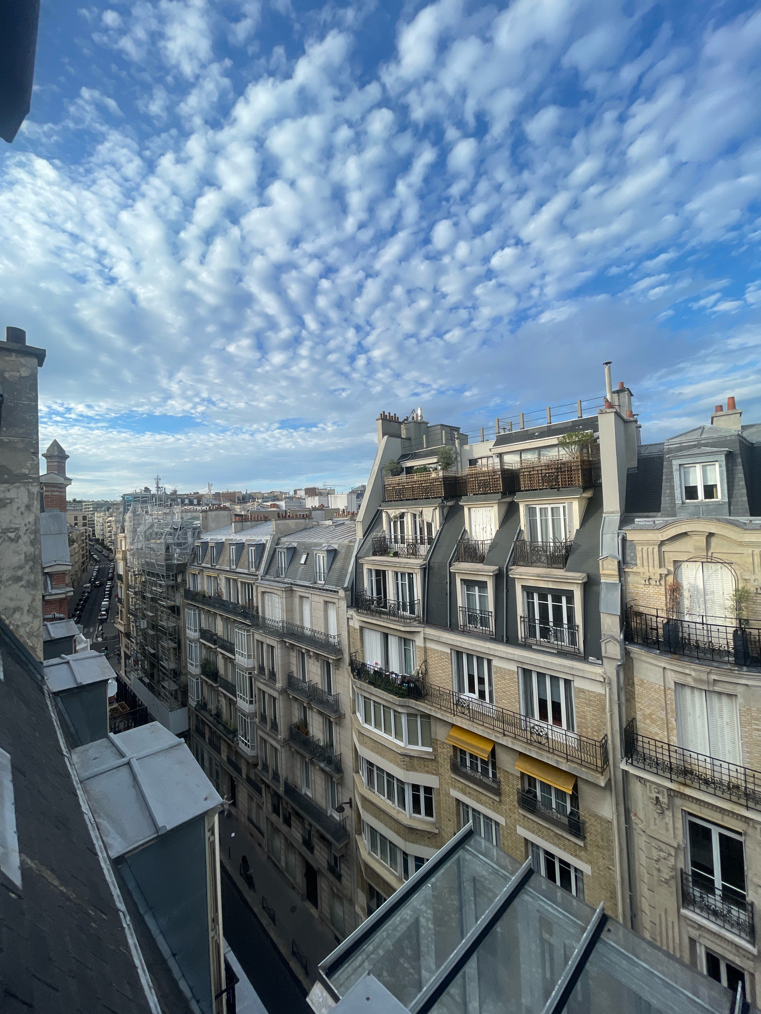 An eye-level, outdoor, full shot captures a panoramic view of Parisian rooftops under a partly cloudy blue sky. The scene overlooks a street lined with Haussmannian buildings, characterized by their cream-colored stone facades, mansard roofs with dormer windows, and intricate iron balconies. Some buildings display yellow awnings and greenery on their balconies. Construction scaffolding is visible on one building in the mid-ground. The foreground features the dark, sloped roof of a building with metallic roof details and a glass structure. The street below is narrow, with parked cars and a few silhouetted pedestrians visible. The image appears to be taken from a high vantage point, likely a rooftop or a high floor window, looking down and across the urban landscape.