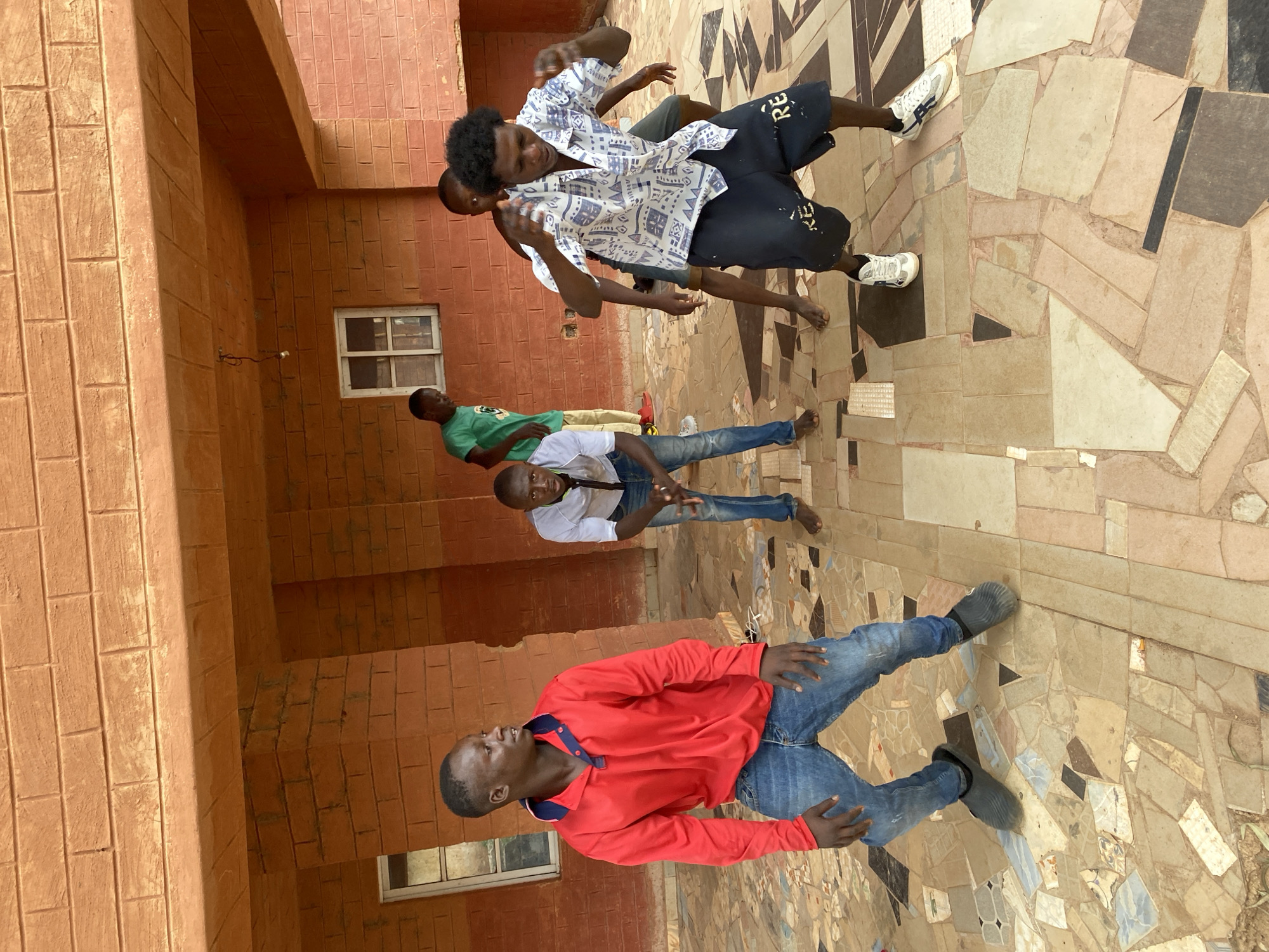 The image, taken in Vom, Nigeria, depicts a group of six young men gathered outdoors during daytime. The scene is set against a reddish-brown brick building and features a strikingly patterned tiled floor.
In the foreground, a man in a bright red long-sleeved polo shirt and blue jeans stands with his hands slightly raised, looking towards the left side of the frame. Behind him, another man in a white short-sleeved shirt and blue jeans looks over his shoulder towards the camera, his left hand slightly open. Adjacent to them, a man wearing a green t-shirt and light-colored trousers is partly visible.
Further back and to the right, two men are in more active stances. One wears a distinctive white short-sleeved shirt with a black pattern and black shorts that display partial text "RE" and "KE". His hands are raised and slightly bent, and he has a focused expression. Another man is partially obscured behind him, also wearing dark shorts. One more young man is visible standing barefoot in this area.
The background consists of a building wall made of reddish-brown rectangular bricks or tiles. A white-framed window is visible on this wall. The floor beneath the men is covered in a complex mosaic pattern of irregularly shaped tiles in varying shades of beige, brown, black, and white, creating a visually interesting ground surface. The overall impression is a casual gathering of friends in a distinctive architectural setting.