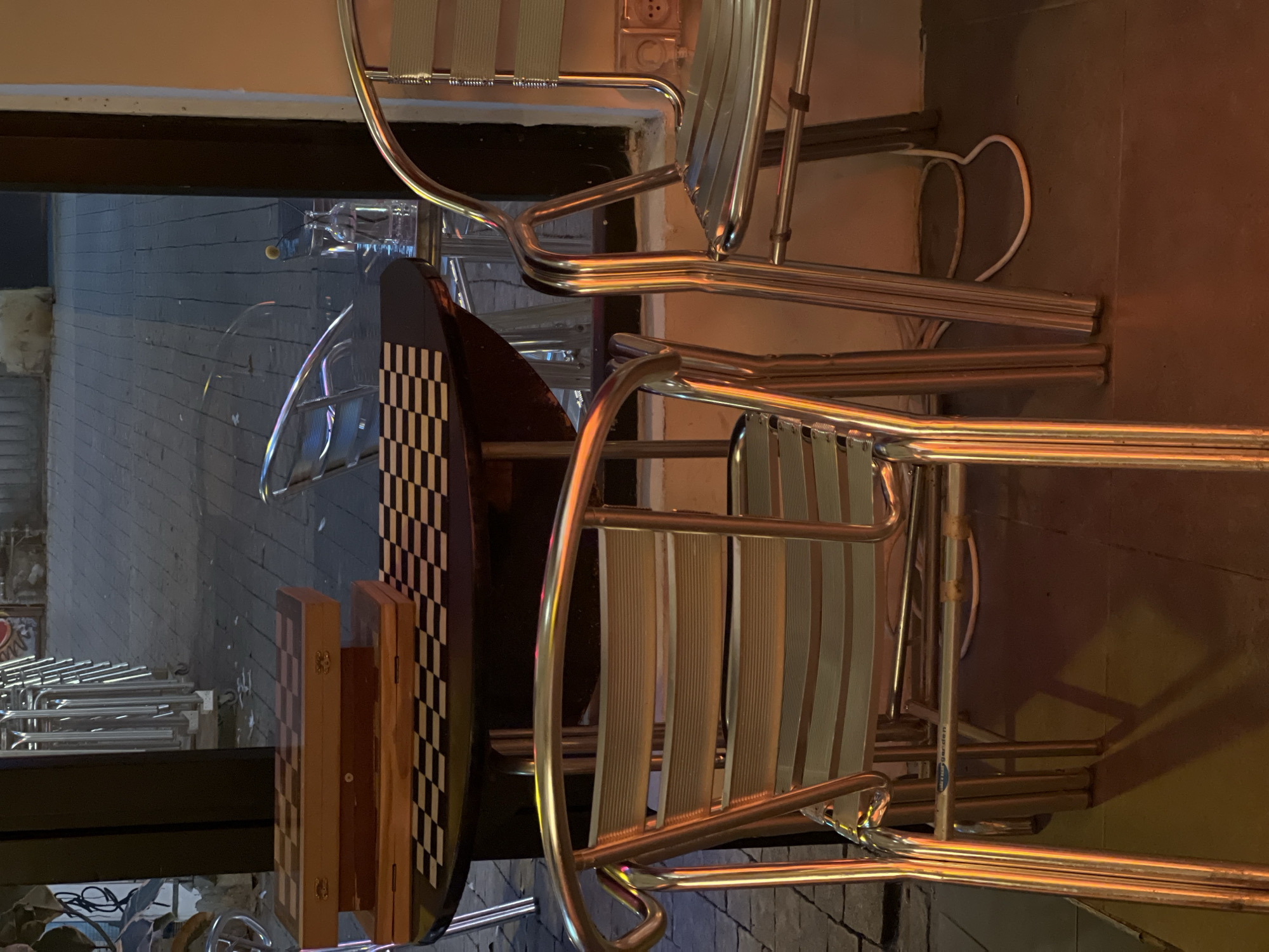 The image shows a cafe setting in Tel-Aviv, Israel. The outdoor area features a small table with a checkerboard pattern and three stacked chrome metal chairs with woven seats. The foreground is illuminated by artificial lighting. Through the open doorway and the transparent chairs, the street is visible, with a pale blue brick surface. An electrical outlet is visible on the wall. There are no people visible in the scene.