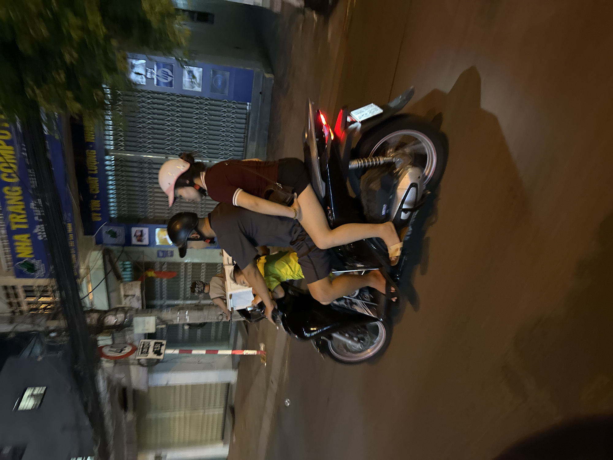Here is a description of the image:
Two people are riding a motorbike on a street in Bac Nha Trang Ward, Vietnam. The person driving is wearing a dark shirt, dark shorts, and a helmet. The person in the back is wearing a pink helmet, a maroon shirt, and dark shorts. The motorbike is black. The street is paved and there are buildings on either side of the road. Overhead electrical wires and utility poles line the street. Signs in Vietnamese are visible on the buildings.