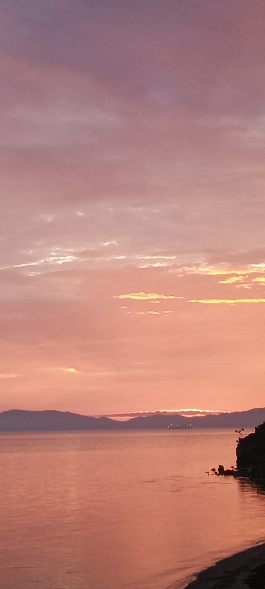 This is a vertical, full-frame, eye-level shot of a sunset over a body of water, likely the Sea of Japan, near Bolshoy Kamen, Russia. The sky is filled with soft, pastel hues of pink and orange, with wispy clouds catching the last rays of the sun. In the distance, a range of dark, silhouetted hills forms the horizon. A calm, reflective body of water, tinted with the colors of the sunset, occupies the lower half of the image. On the right side, a dark, rocky shoreline juts out into the water, with a small patch of sandy beach visible at the very bottom. A distant ship with lights is visible on the horizon between the hills. No people are visible in the scene.