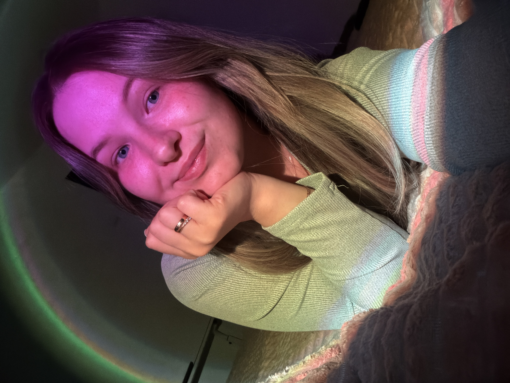 A young woman lies on a fluffy bed. Her head rests on her hand, which has a gold ring on it. She is wearing a green sweater. The background is dark, but a rainbow light shines on the wall behind her and a pink light shines on her face. The location cannot be determined from the image.