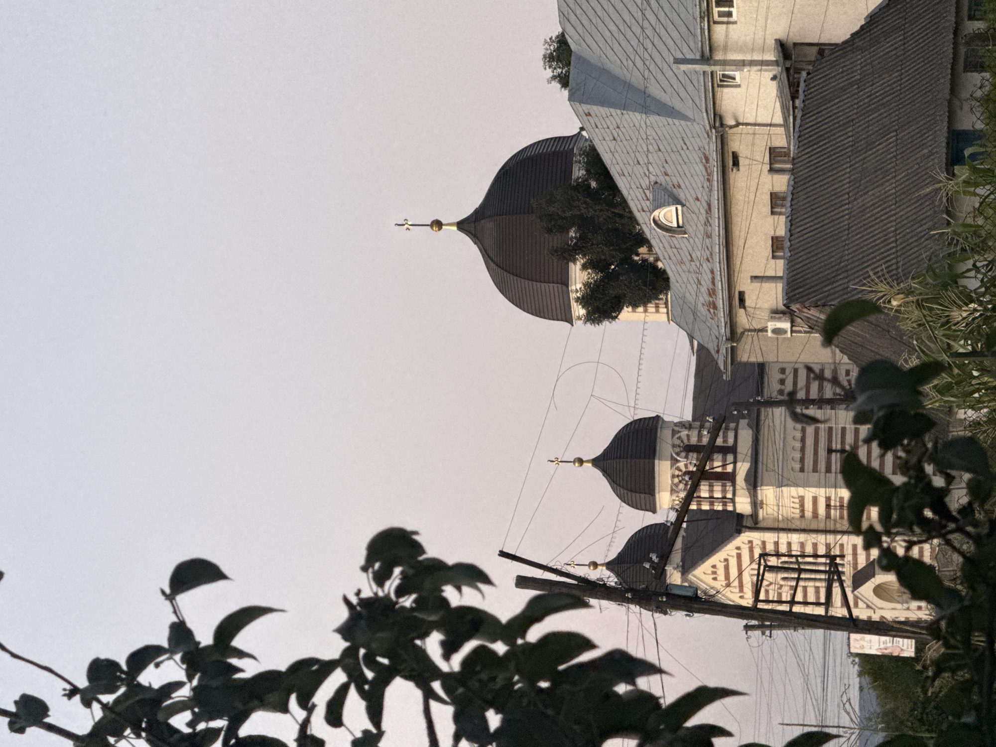 An exterior, low-angle, medium shot captures a view of a church and surrounding buildings, partially obscured by tree branches in the foreground. The sky is a pale, hazy white. The church, with its white and red striped facade, features two dark, domed roofs topped with crosses. To the right of the church, a building with a grey, corrugated metal roof is visible. Another building with a lighter colored facade and a grey pitched roof is situated in front of the first building. Various utility poles and wires are strung across the scene. The overall scene appears to be within an urban or semi-urban environment.