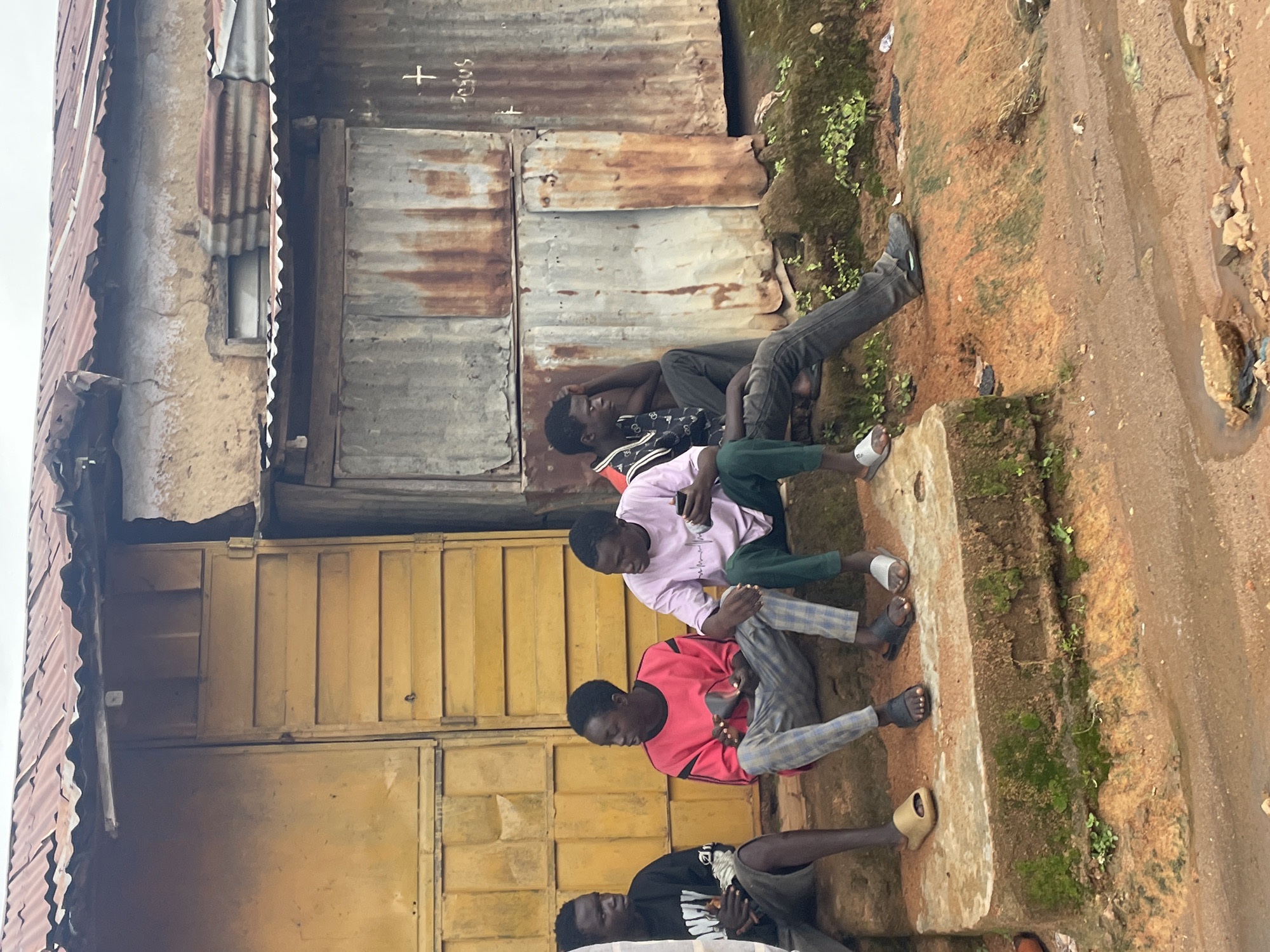 Four young men are sitting on a concrete step in front of a weathered building with a corrugated iron roof and walls. The building has a large yellow door with horizontal slatted panels. On the right side of the building, the corrugated metal walls are rusty and appear to have a white cross and the word "JESUS" scrawled on them. The ground in front of the building is a reddish-brown dirt with some puddles and debris. The men are dressed casually. The man on the far left wears a black t-shirt and shorts. The man next to him wears a bright pink long-sleeved shirt and checkered pants. The third man from the left is wearing a light purple t-shirt and dark green pants, and is holding a smartphone. The man on the far right is wearing a dark patterned shirt and dark pants, and has his hand on his head. They appear to be interacting with each other and looking in various directions. The scene suggests a street-level view in a developing urban area. The location is identified as Jos, Nigeria, which is known for its diverse urban landscape and informal settlements.